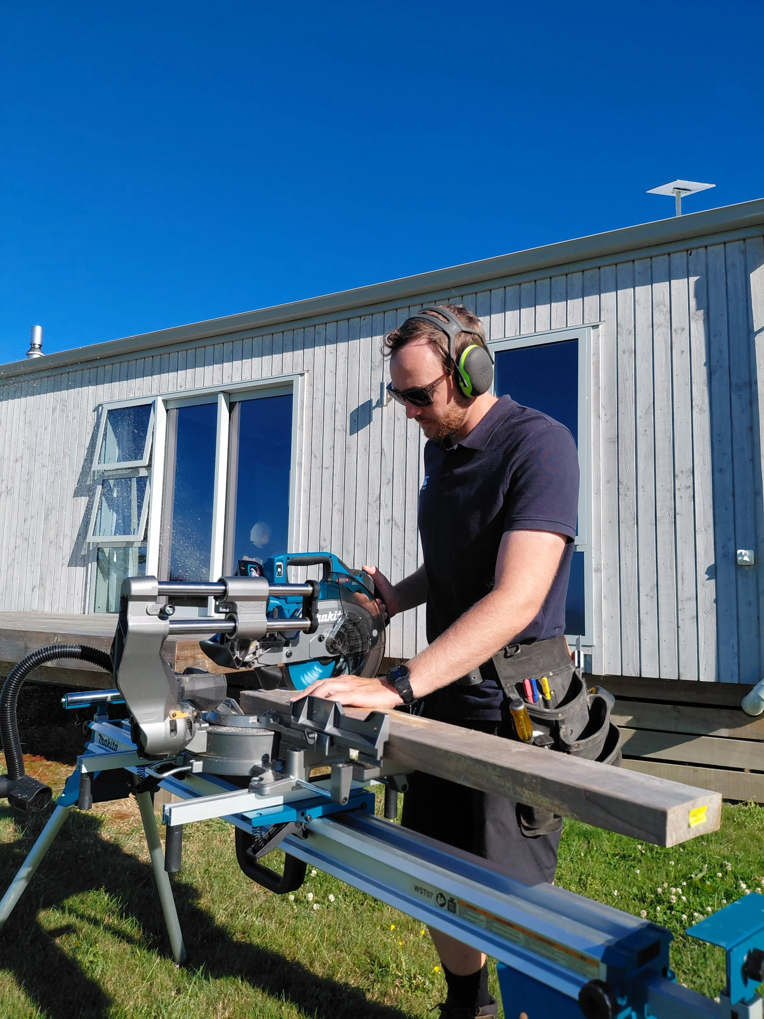 A man wearing safety headphones and sunglasses operates a miter saw outdoors near a modern house with large windows and a blue sky.