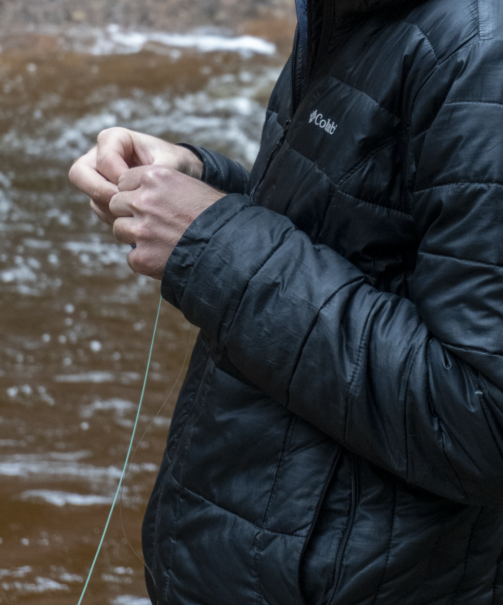 Person wearing a black Columbia jacket, standing outdoors near a body of water with small waves, holding a fishing line.