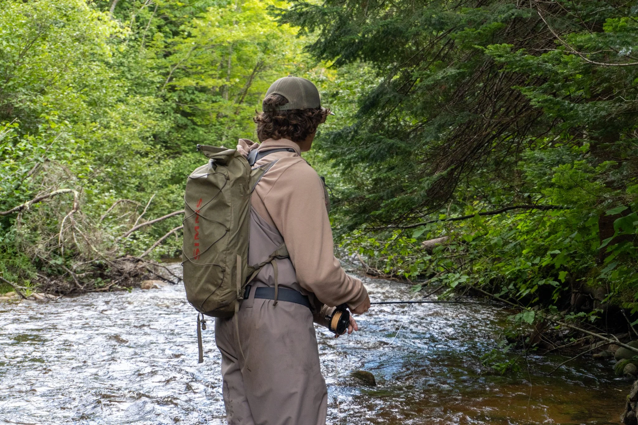 Person with curly hair wearing a hat and outdoor gear fishing in a stream surrounded by green trees.