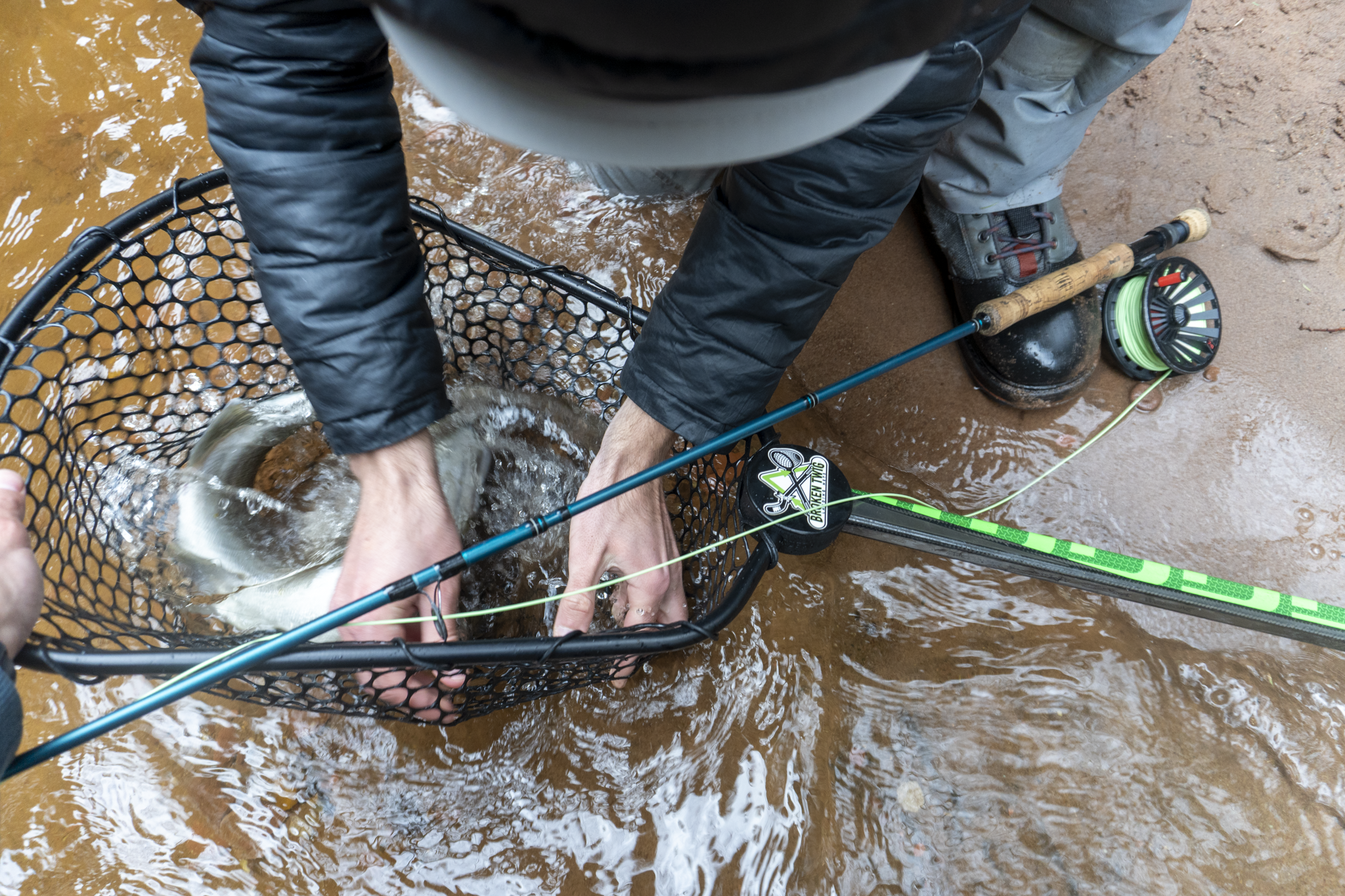 Person wearing a rain jacket and boots, using a fishing net to catch a fish in a muddy river with a fishing rod nearby.