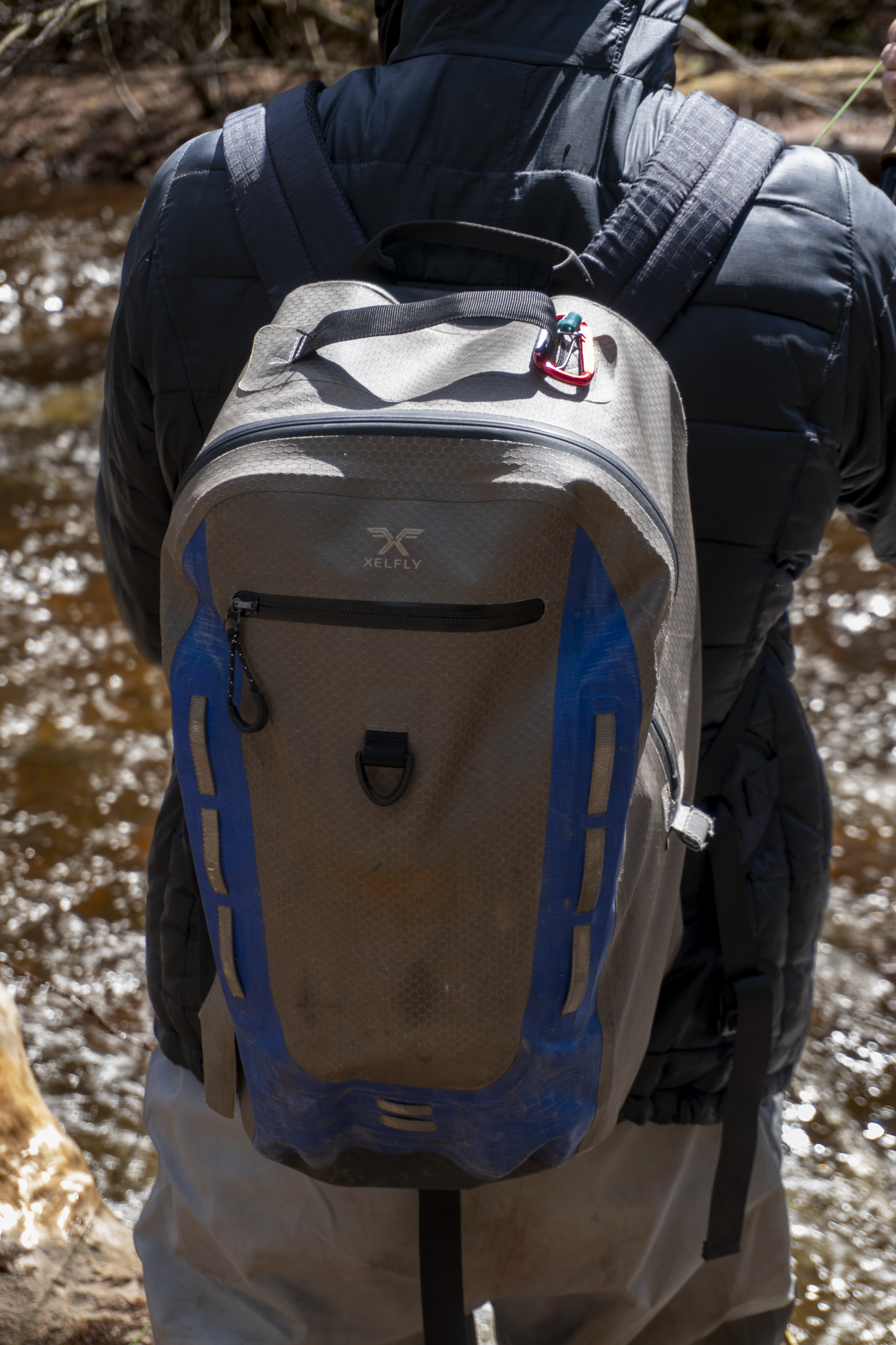 Person wearing a jacket and a backpack with a carabiner clipping a small item onto it, standing near a stream or river.