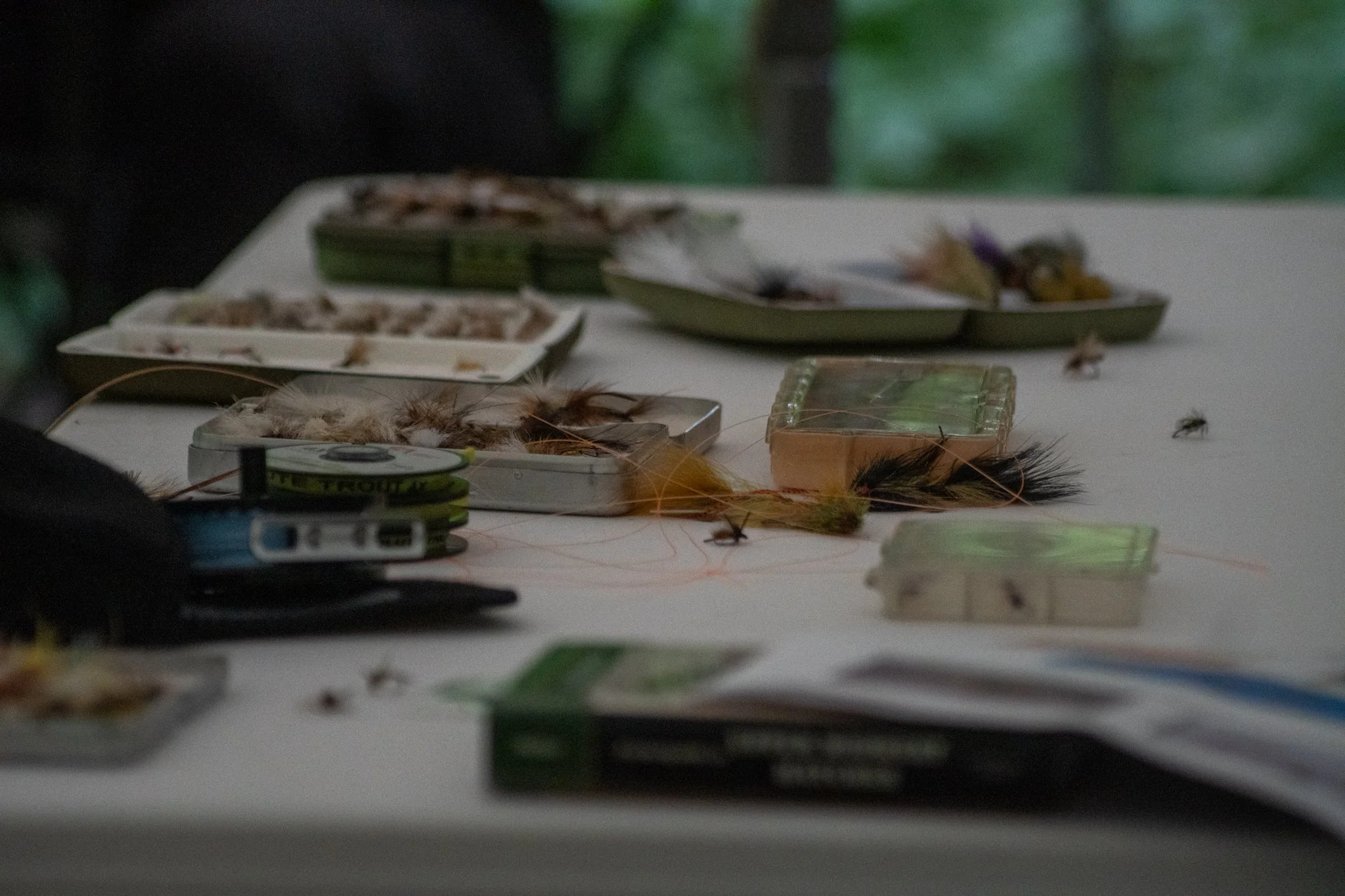 Table with various fishing flies and tackle, including boxes of artificial flies, some feathers, and a fishing reel.