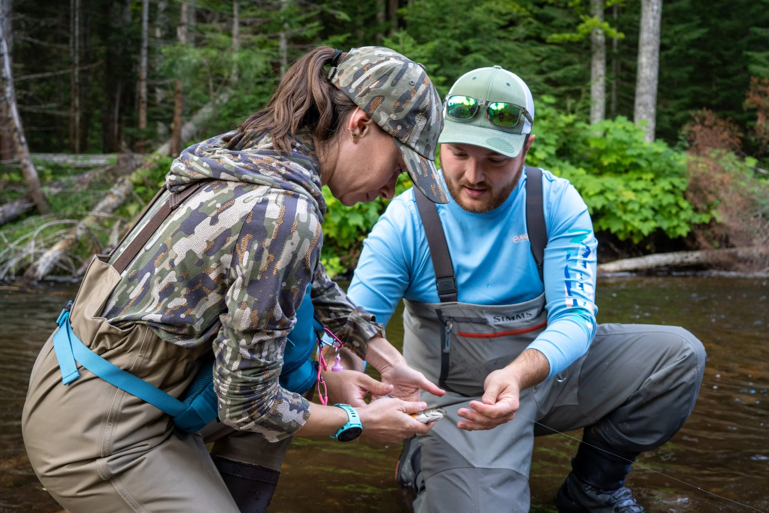 Two people outdoors near a riverbank, one woman and one man, examining a small fish together wearing outdoor gear and baseball caps, surrounded by green trees.