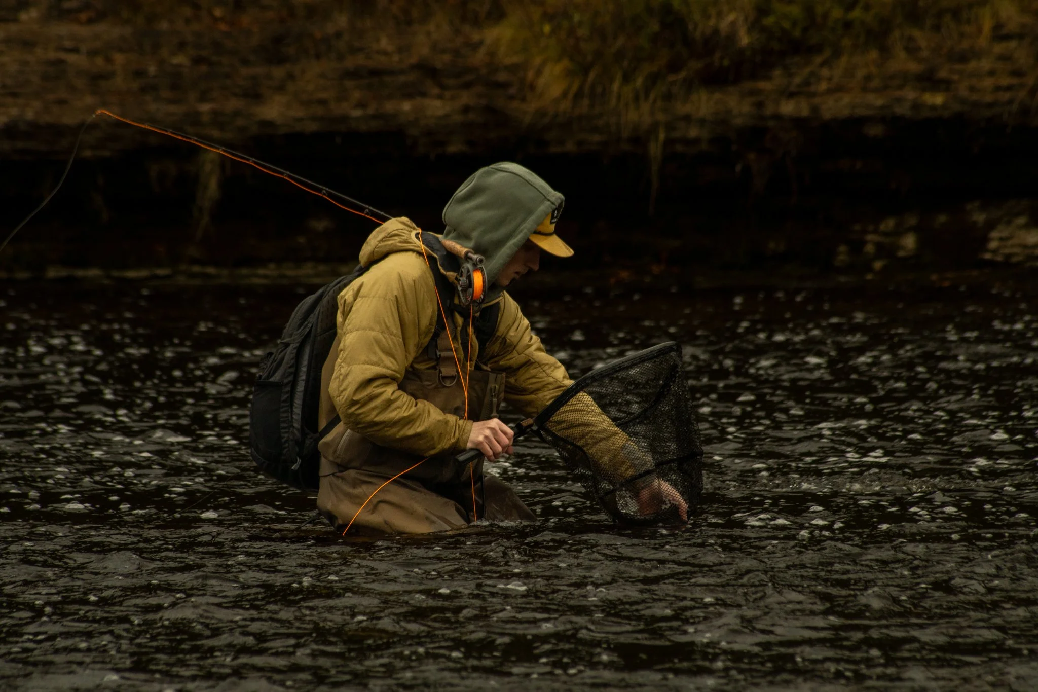 A person wearing outdoor gear, including a hoodie, backpack, and headphones, stands in a dark, shallow river or lake, holding a fishing net and preparing to fish.