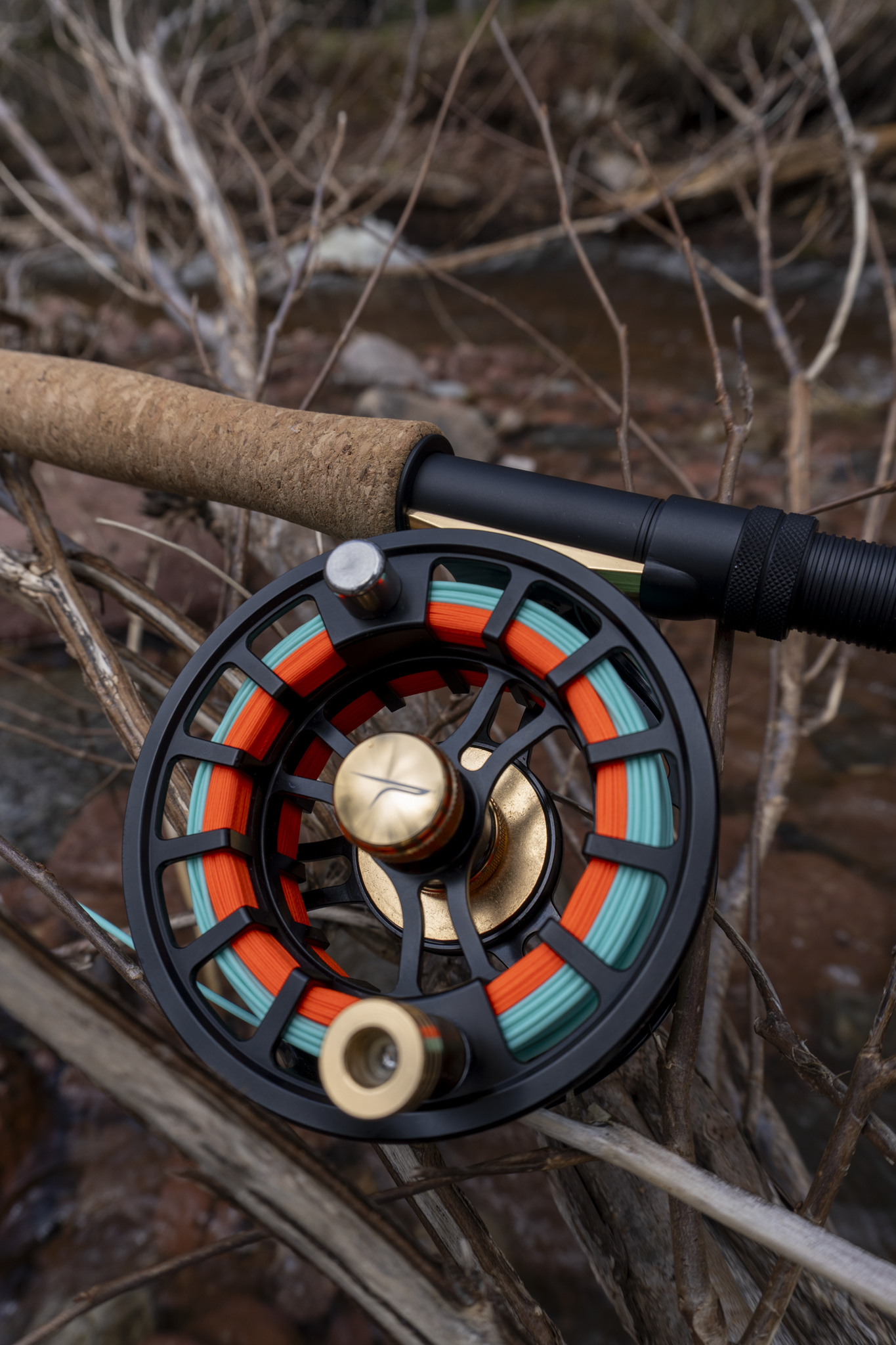 A close-up of a fly fishing reel with an orange and blue line, resting on dry twigs and branches outdoors.