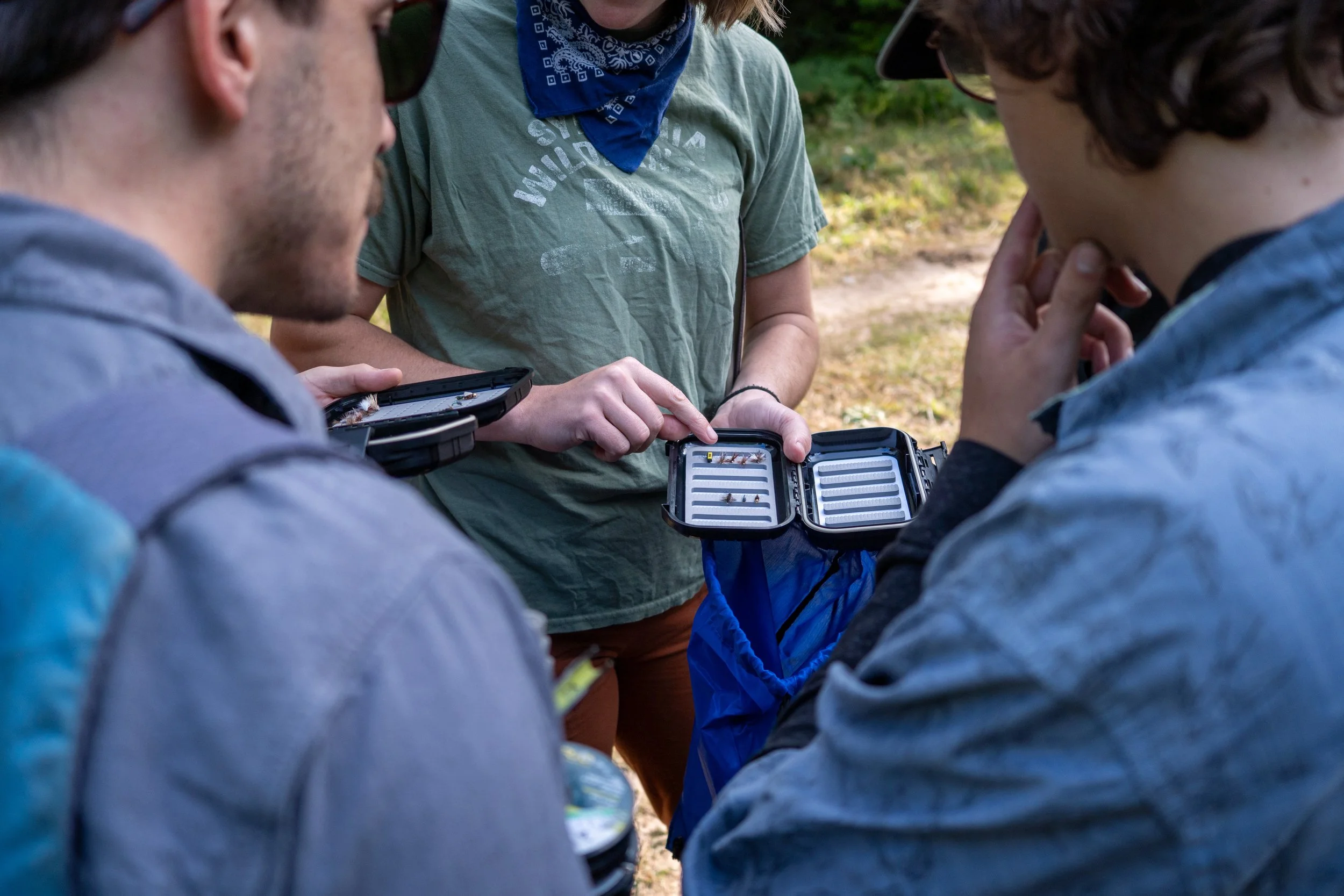 Three people outdoors looking at an open fly book, with one person holding the case containing flies.