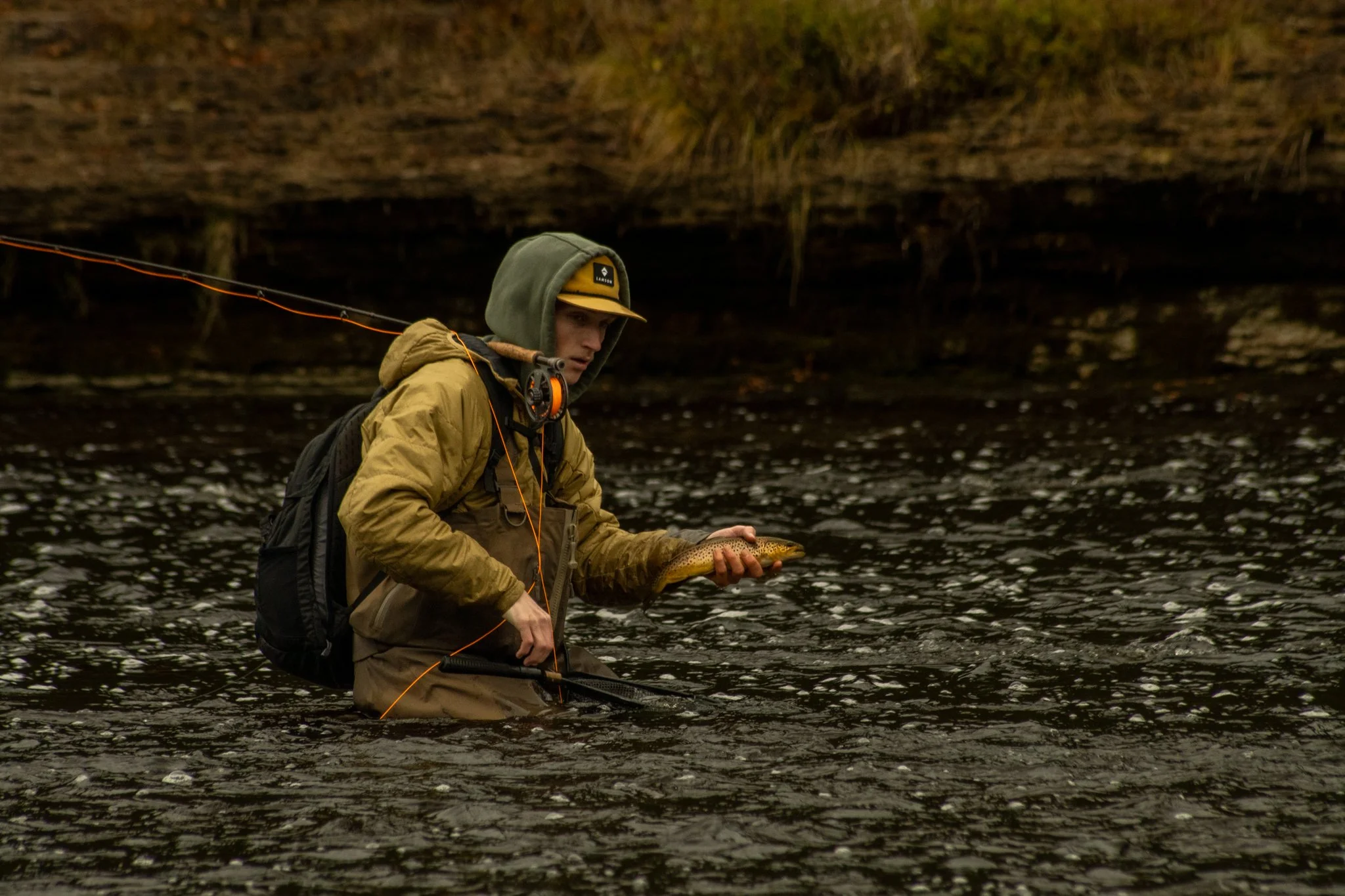 A person wearing outdoor gear, including a tan jacket, green hood, and a yellowish cap, standing in a river and holding a fish.