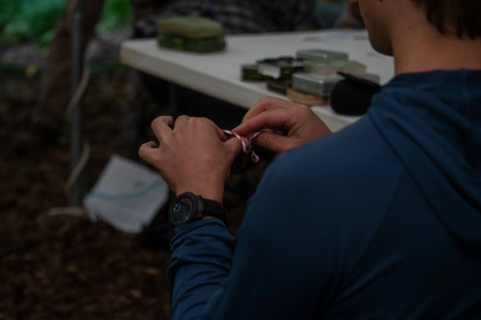 A person with a blue jacket and black watch knotting a colorful rope, with a table of various small packs or containers of supplies in the background, outdoors.