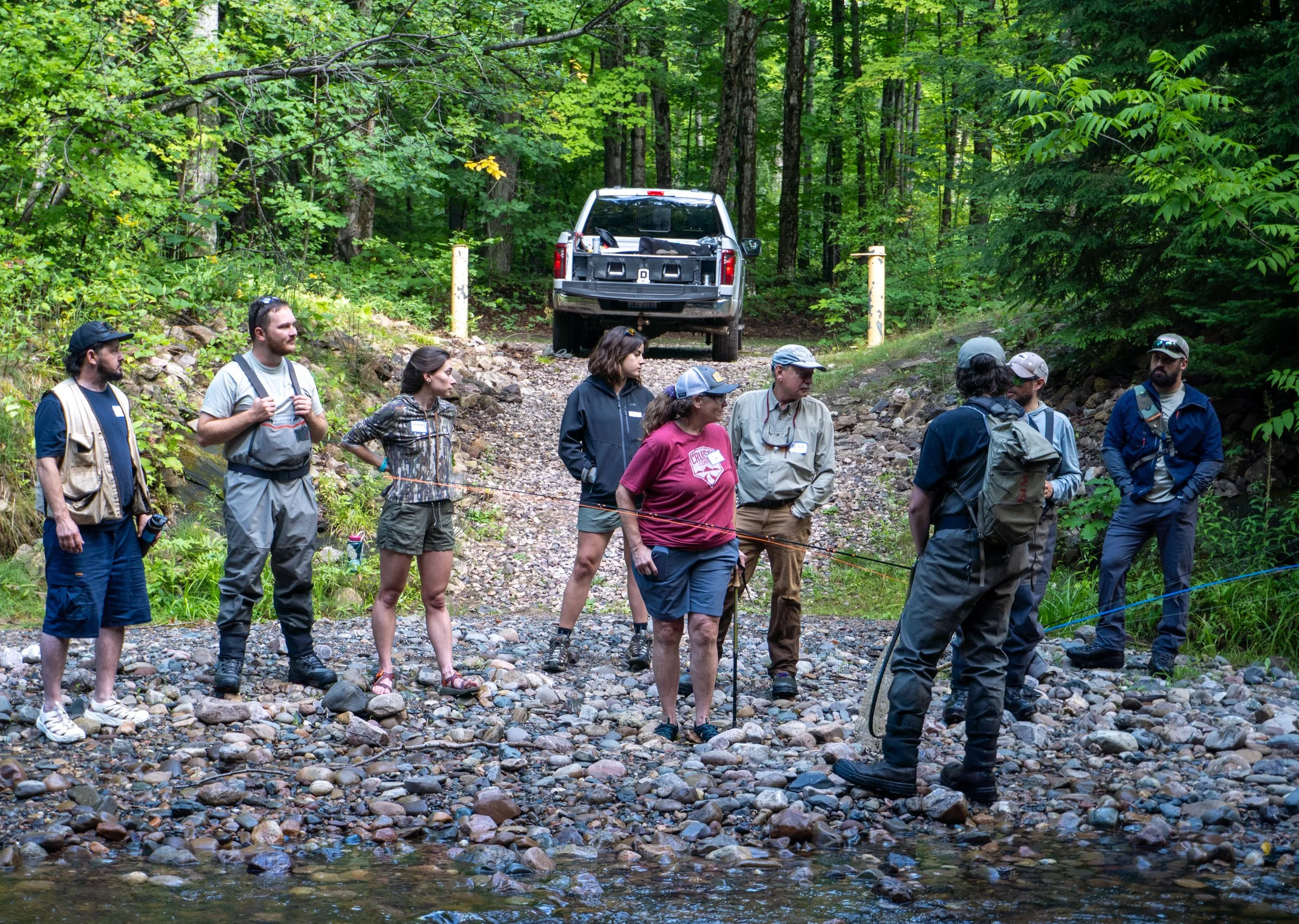 A group of people, mostly wearing outdoor clothing and hats, standing on a rocky creek bed in a dense green forest. There is a pickup truck parked in the background among the trees.