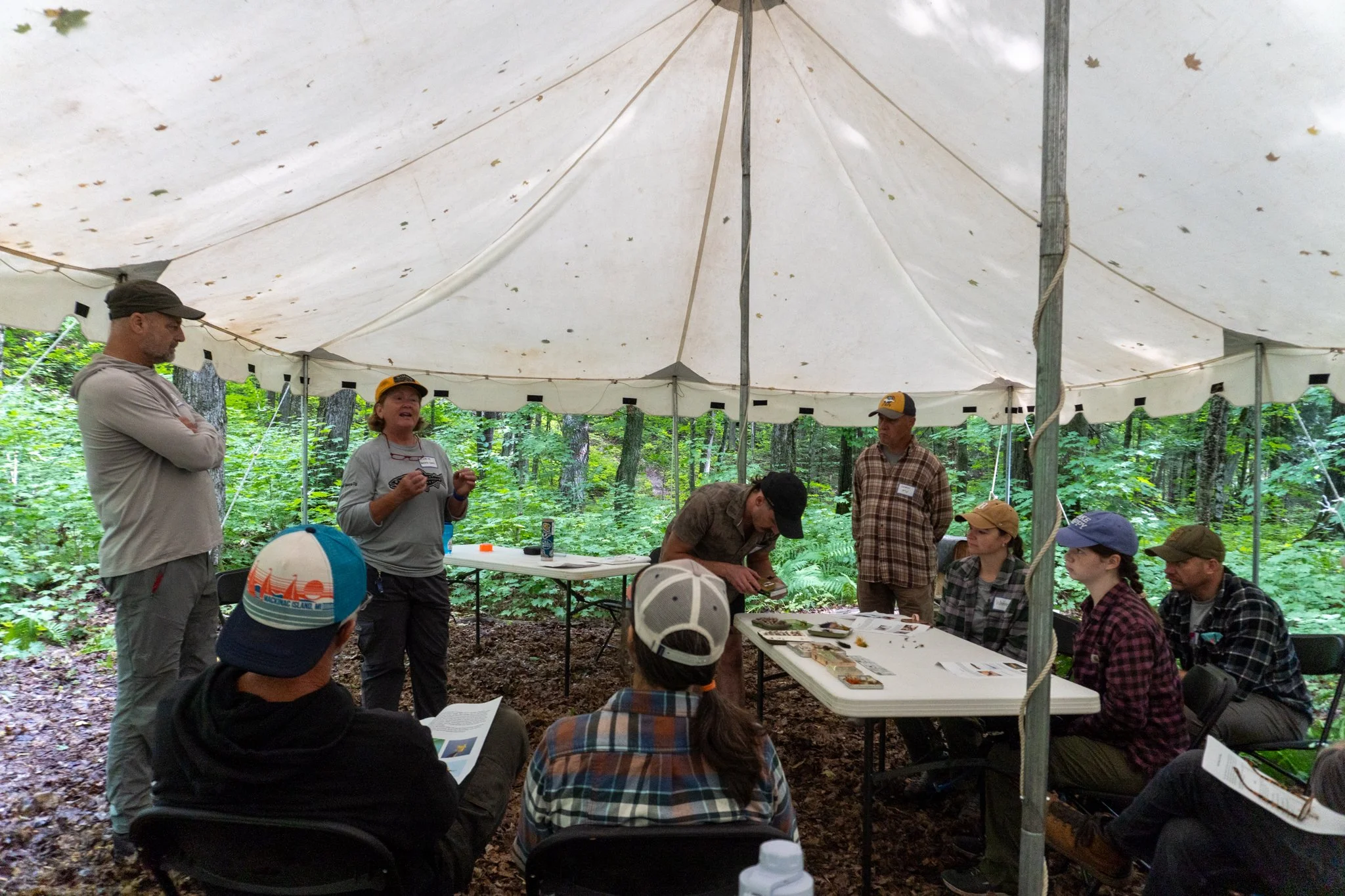 A group of people under a white canopy tent in a wooded area, engaged in a discussion or presentation. Some are standing, others sitting, with tables and papers visible. The scene appears to be an outdoor event or meeting.