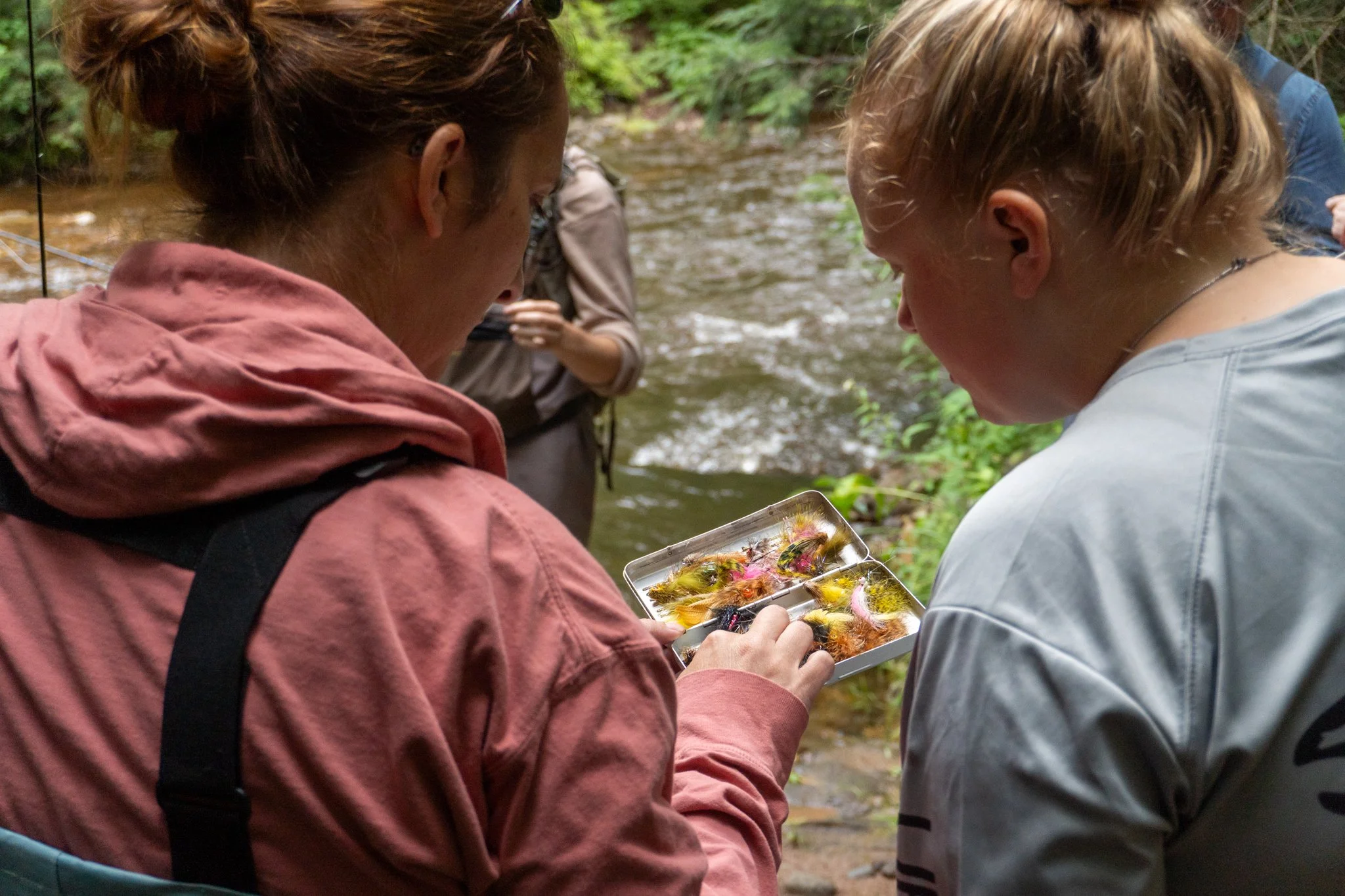 Two people examine a plastic container filled with colorful fishing flies outdoors near a stream in a forest.