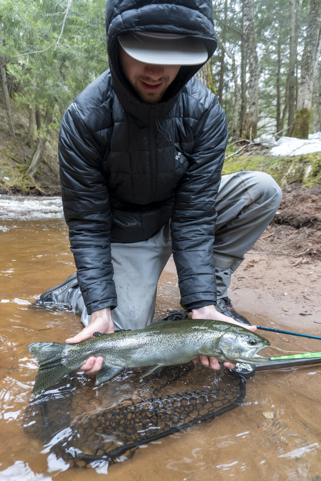 A man in outdoor clothing kneels in a river while holding a large fish he just caught, with trees and snow in the background.