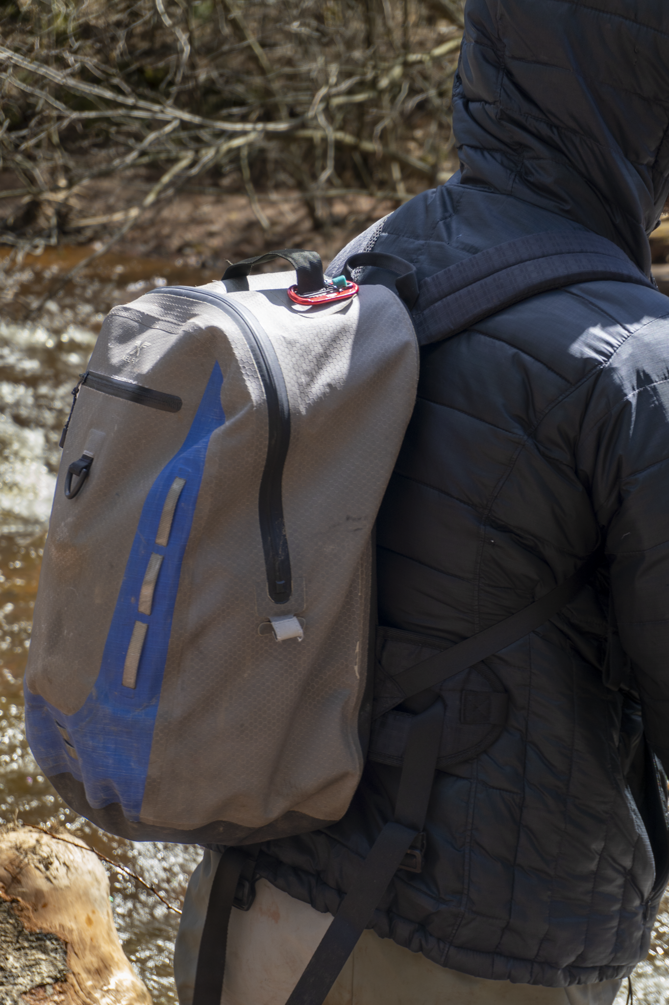 Person wearing a black jacket and gray pants, carrying a gray backpack with blue accents, hiking outdoors near a stream with rocks and leafless branches in the background.