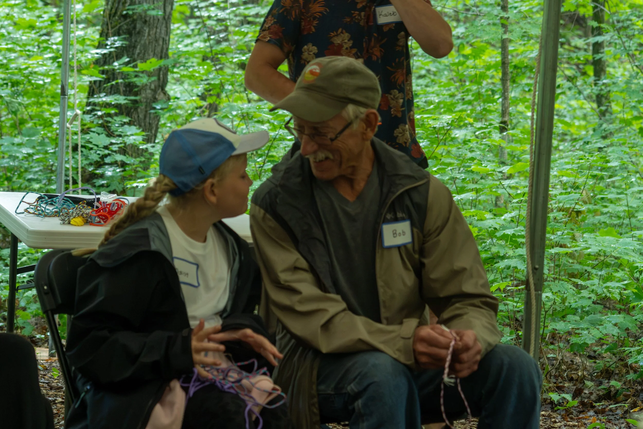 An elderly man with glasses and a tan cap, holding a pink rope, sitting and talking with a young girl wearing a cap and a black jacket, in a wooded outdoor setting with green foliage in the background. Someone is standing behind them, partially visib