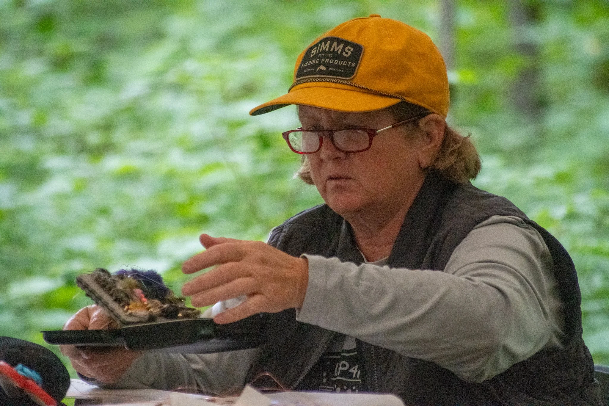 A person wearing a yellow cap with a patch reading "Simms Fishing Products" and glasses, focused on handling a butterfly on a tray.