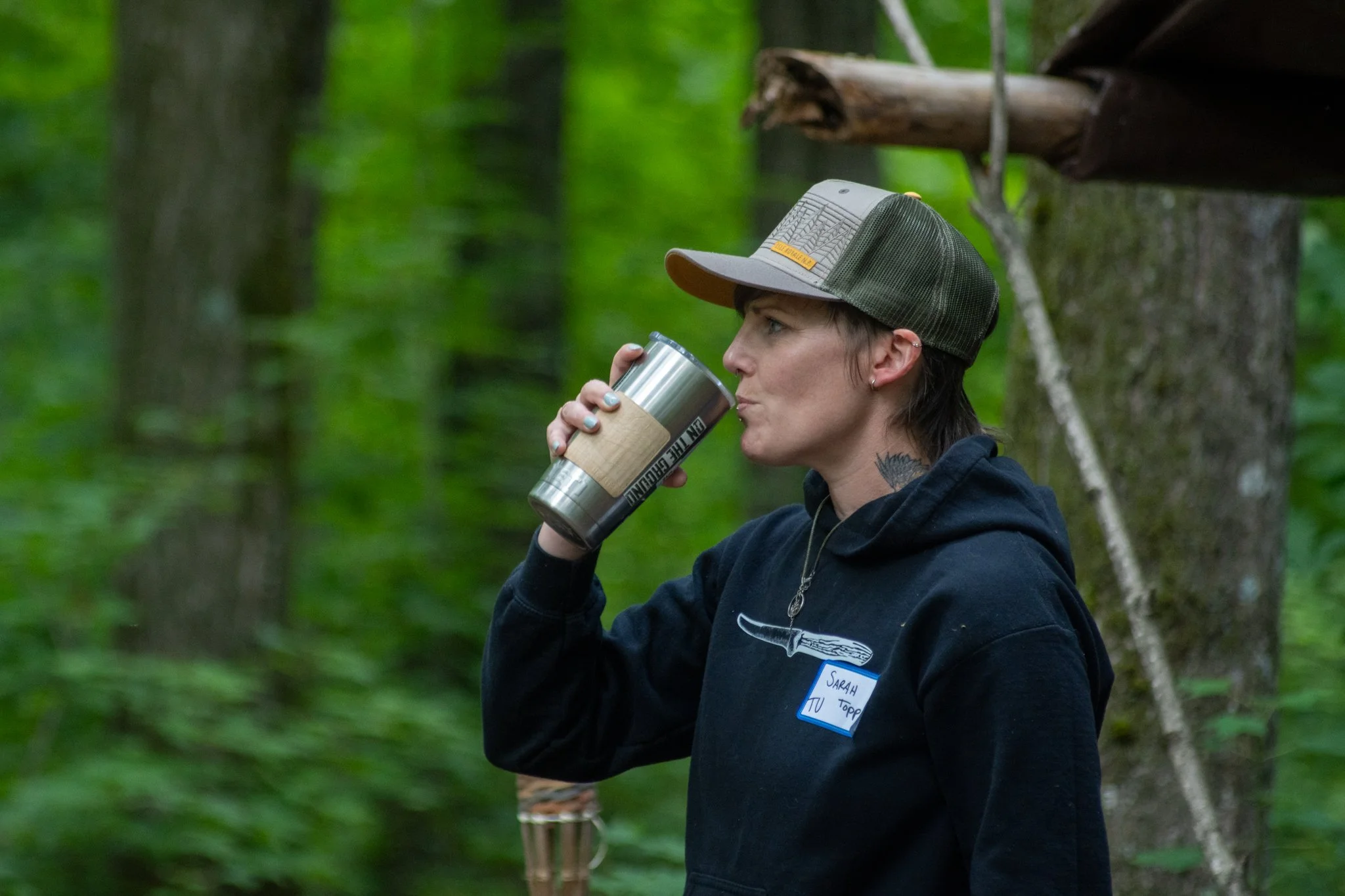 A person wearing a cap drinking from a stainless steel travel mug in a forested area.