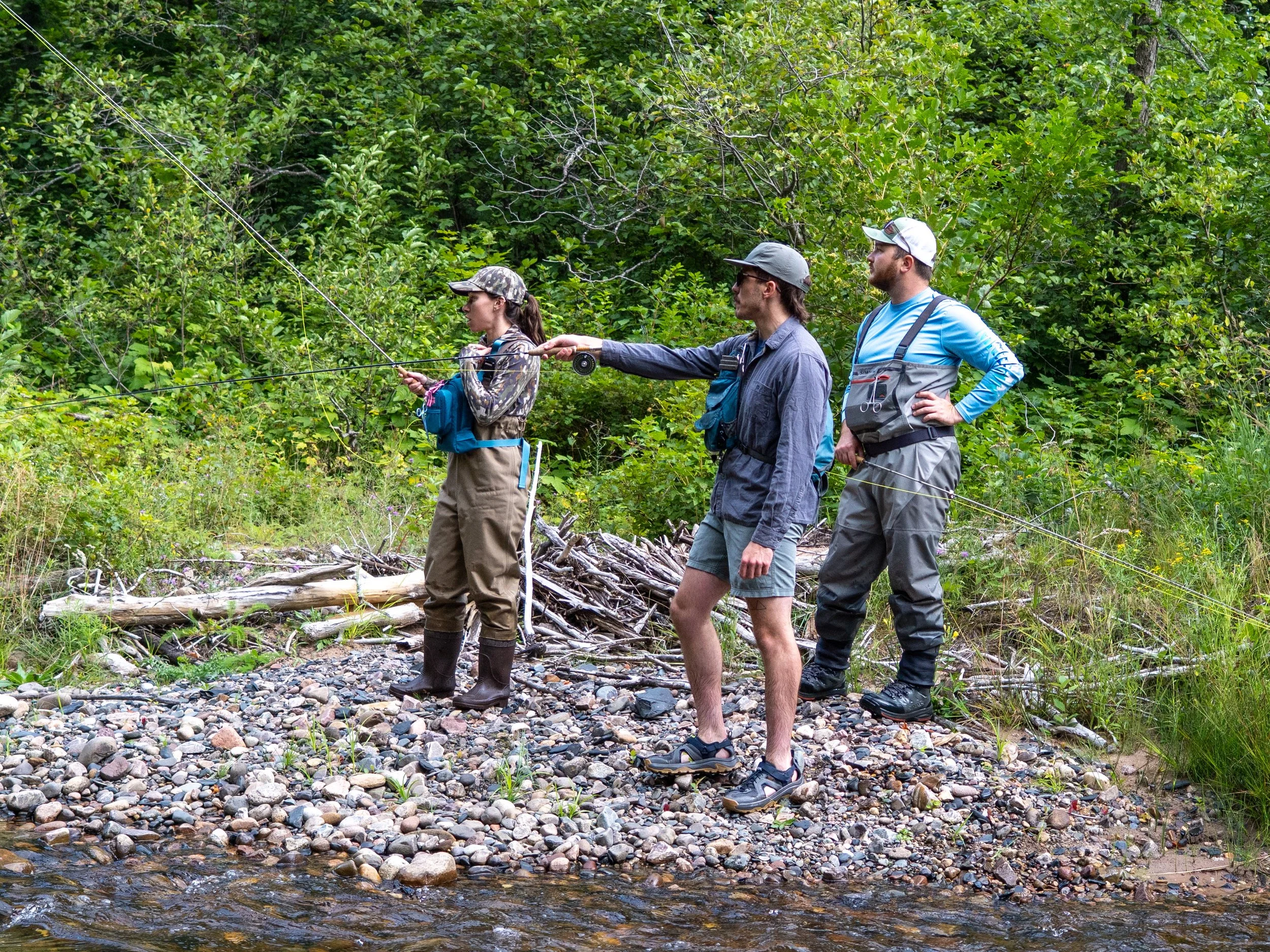 Four people standing on rocks near a small stream in a forest, fishing with a fly rod.