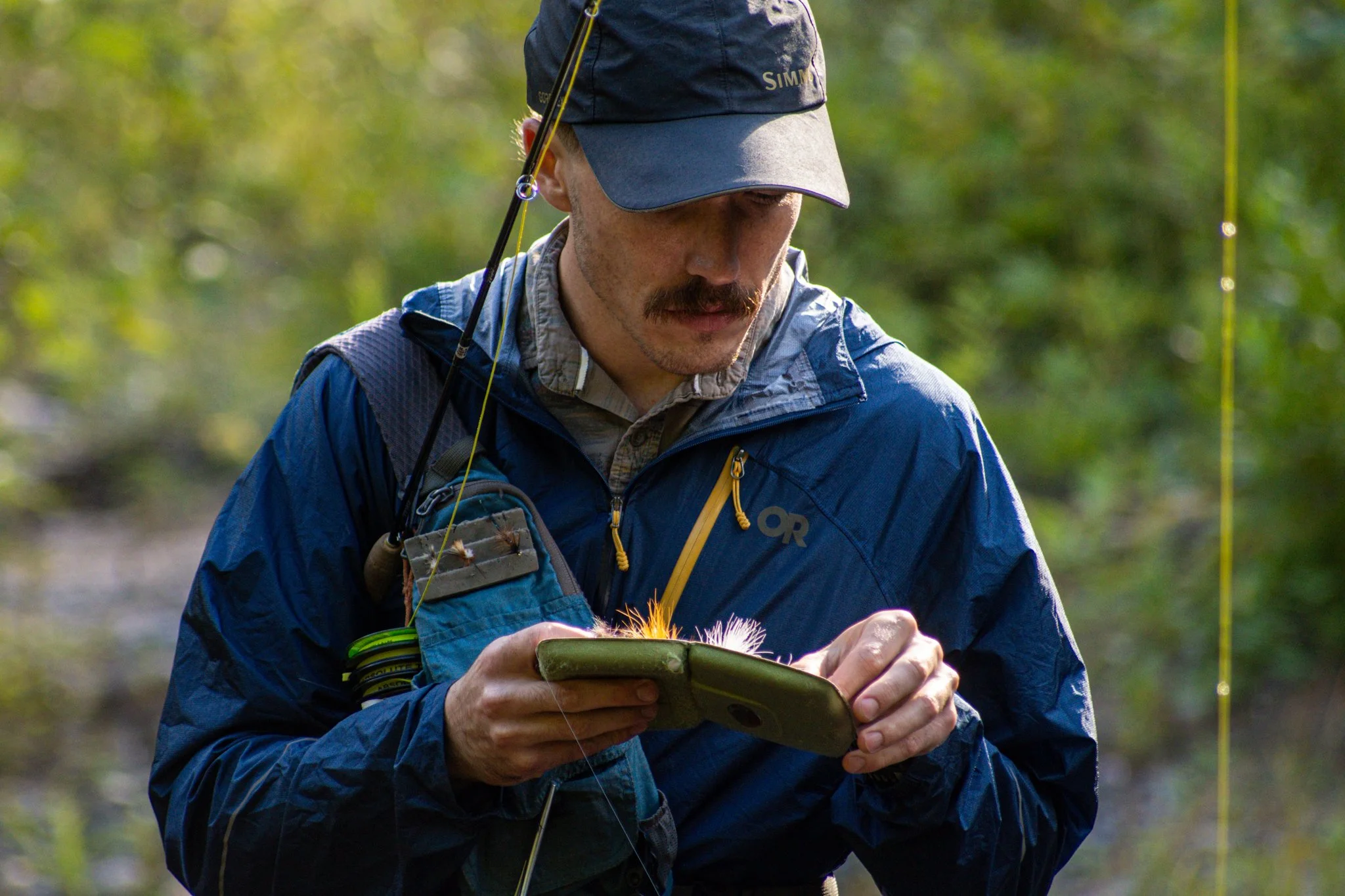A man in outdoor gear, including a blue jacket and gray cap, is looking at a smartphone while holding it with both hands. He is outdoors with blurred greenery in the background.