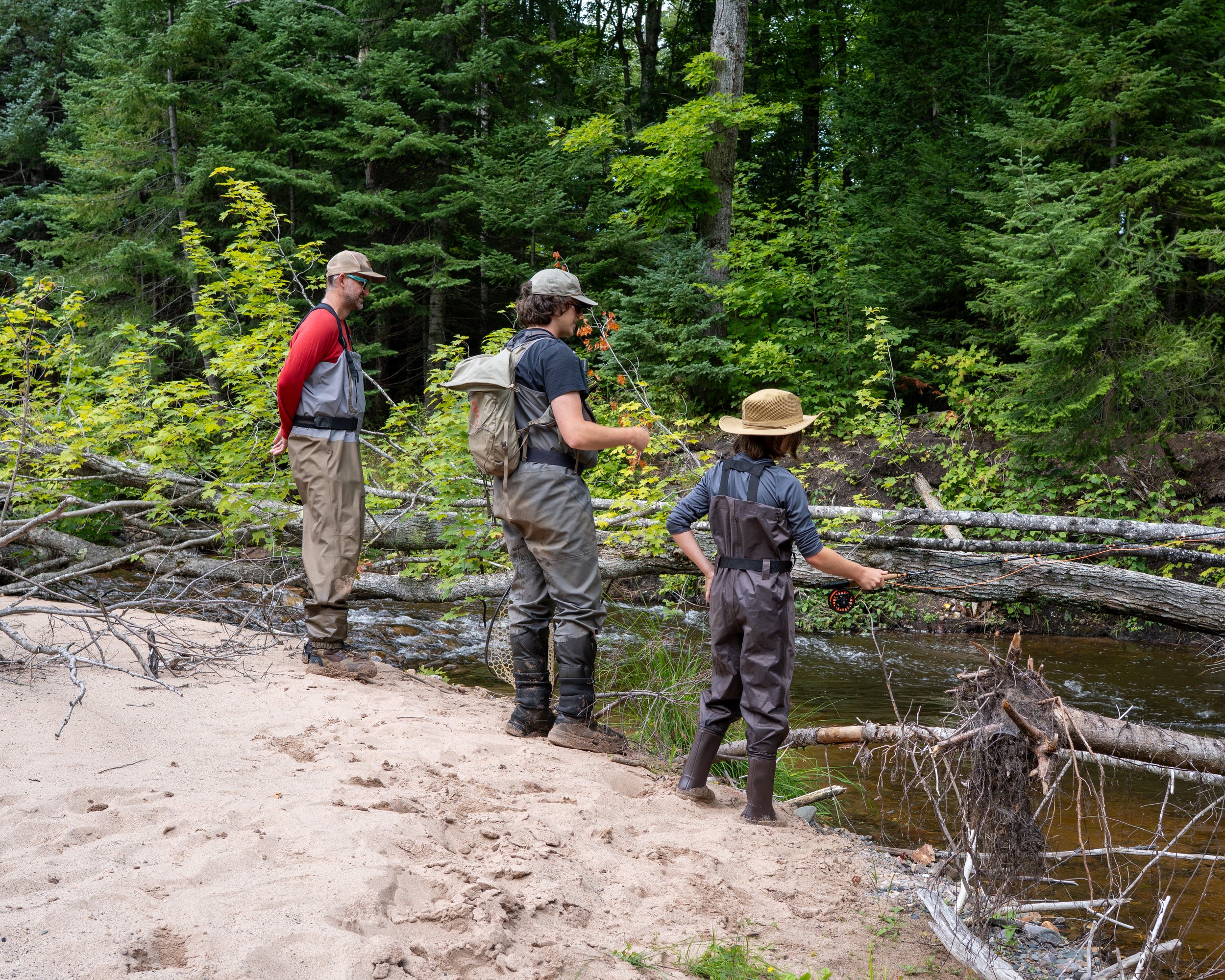 Three people, two adults and one child, stand by a small stream in a lush, green forest. They are dressed in outdoor gear, including hats and backpacks, and fly fishing and enjoying nature.