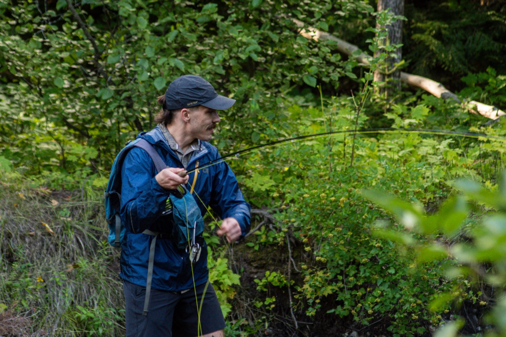 A man fishing in a lush green forest with dense foliage, wearing a blue jacket and a cap.