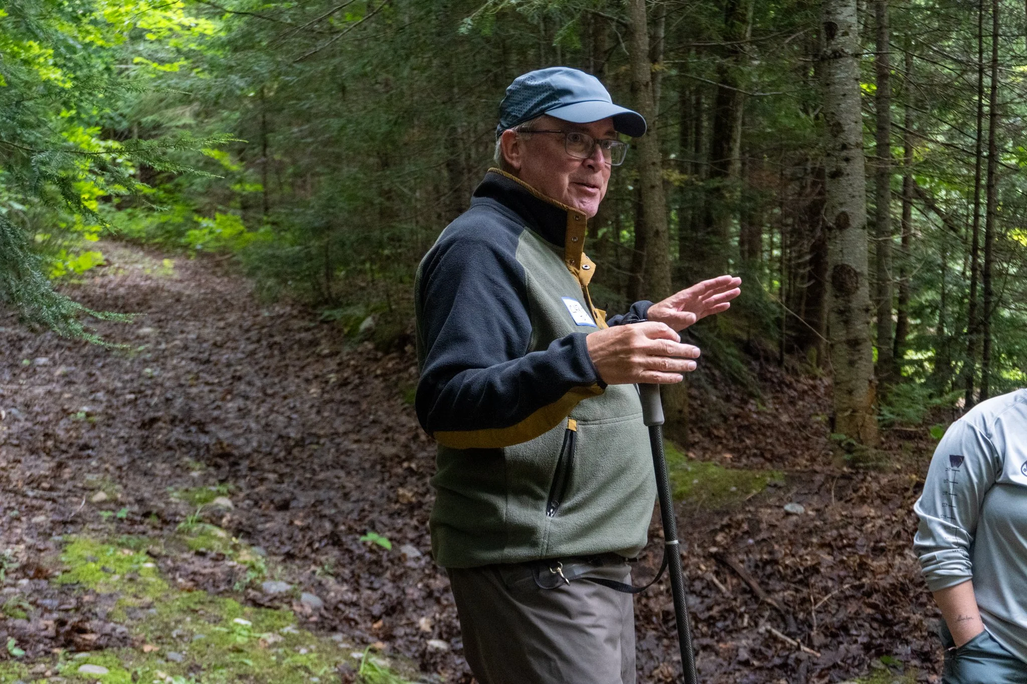 An elderly man wearing glasses, a blue cap, and outdoor clothing, standing in a forest with trees and a dirt path, appearing to be explaining or discussing something.