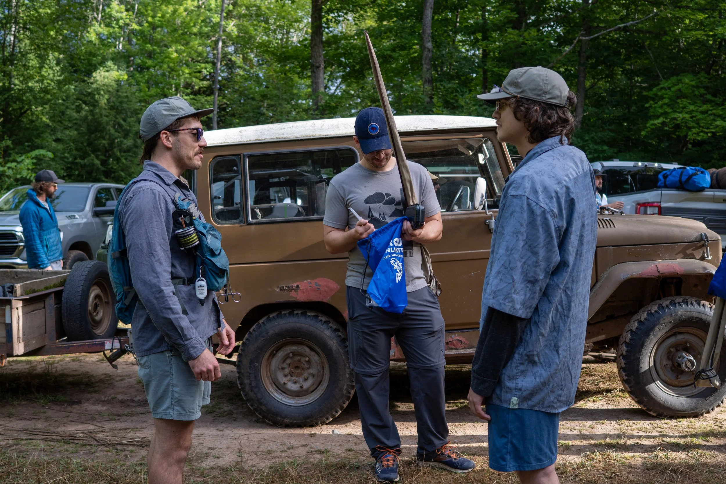 Group of people outdoors near a brown off-road vehicle, with lush green trees in the background, engaged in conversation.