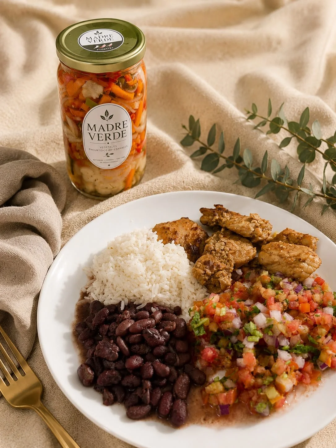 plato con arroz, frijoles negros, pollo con costra de sésamo, y pico de gallo con jitomate, cebolla y chile, acompañado de un frasco de vegetales en vinagre y un tenedor dorado sobre un mantel beige.