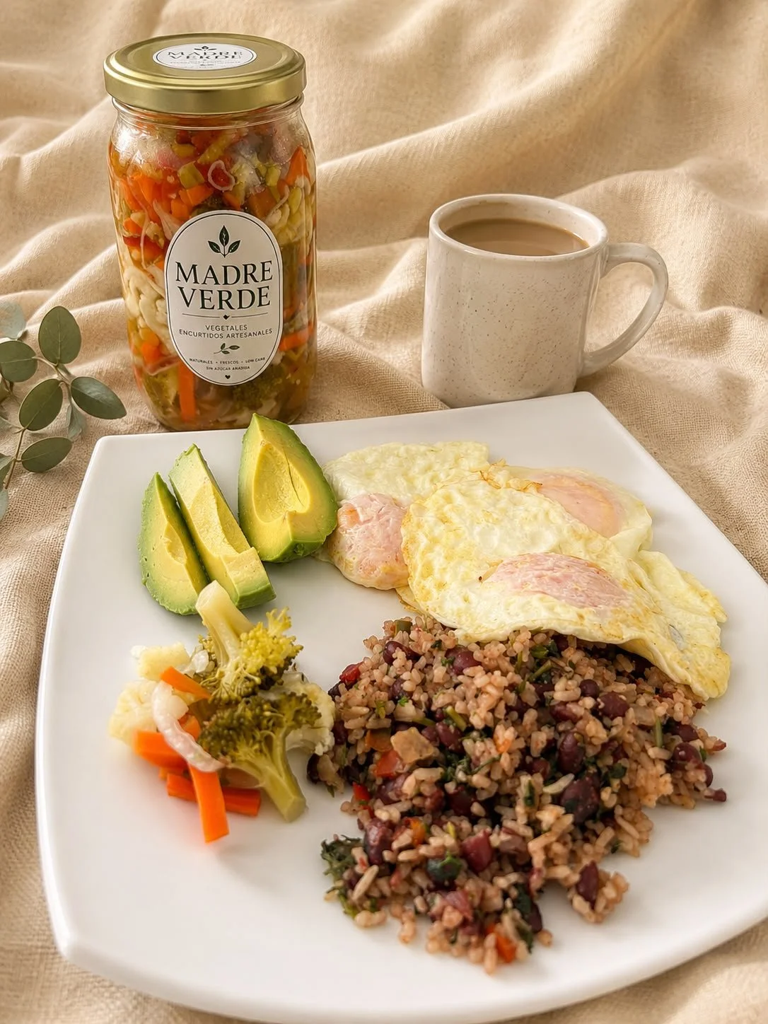 Plato de desayuno con aguacate, huevos fritos, arroz con frijoles, ensalada de verduras, una taza de café y un frasco de vegetales encurtidos.
