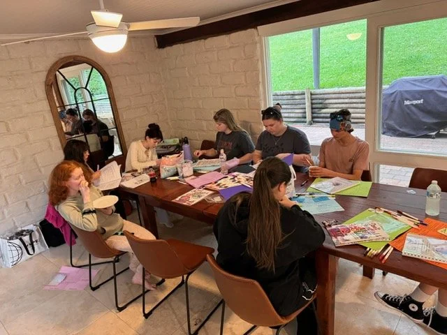 Group of children and teenagers sitting at a large wooden table in a bright room, engaging in arts and crafts activities with various supplies, with a large window showing a green lawn outside.