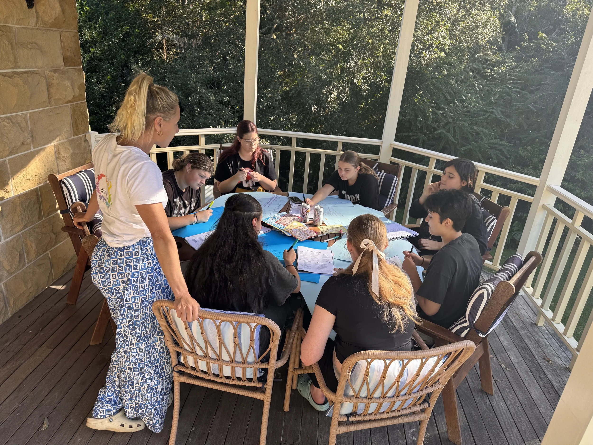 Group of people gathered around a table on an outdoor porch, engaged in a discussion with papers, notebooks, and drinks on the table, surrounded by trees.