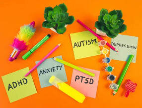 Note cards labeled ADHD, Anxiety, Autism, Depression, and PTSD surrounded by colorful pens, small plants, and decorative items on an orange background.