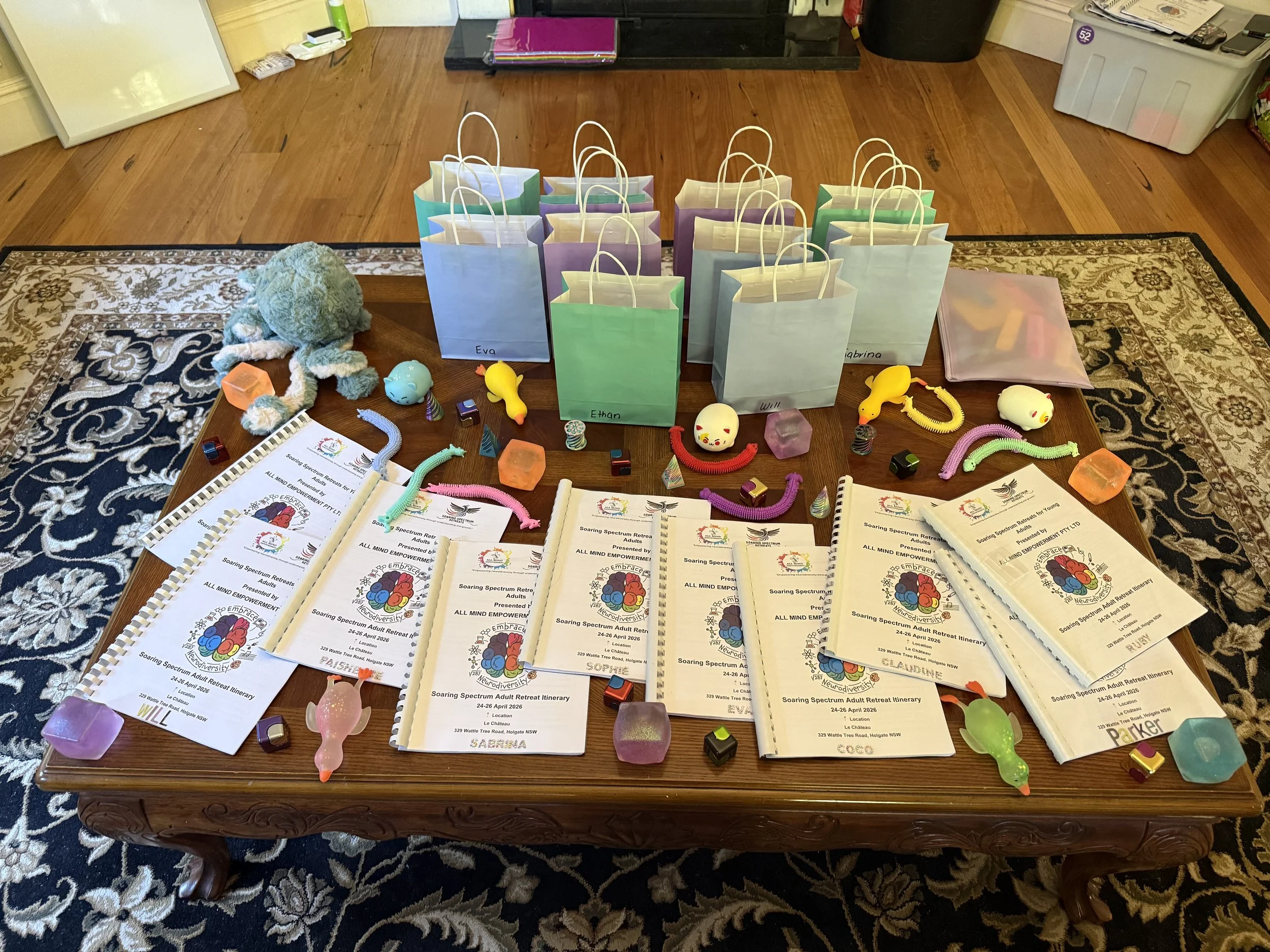 A table with colorful gift bags, some labeled with names, surrounded by small toy figurines, plastic jewelry, and printed retreat program booklets, in a room with a patterned rug and a wooden floor.