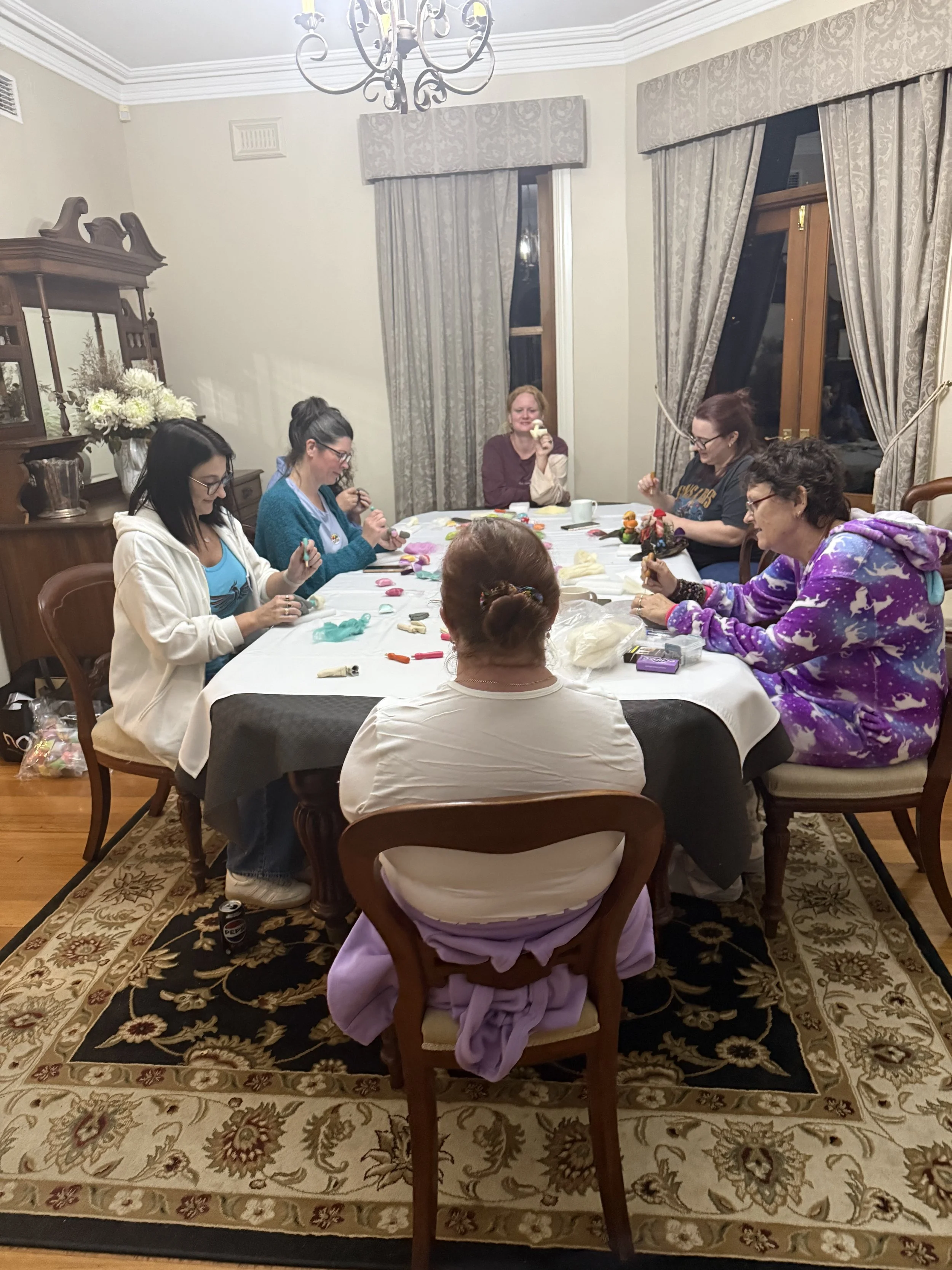 A group of six women sitting around a dining table, working on crafts or sewing projects, with crafting supplies on the table in a well-lit, decorated room with floral curtains and a wooden sideboard.