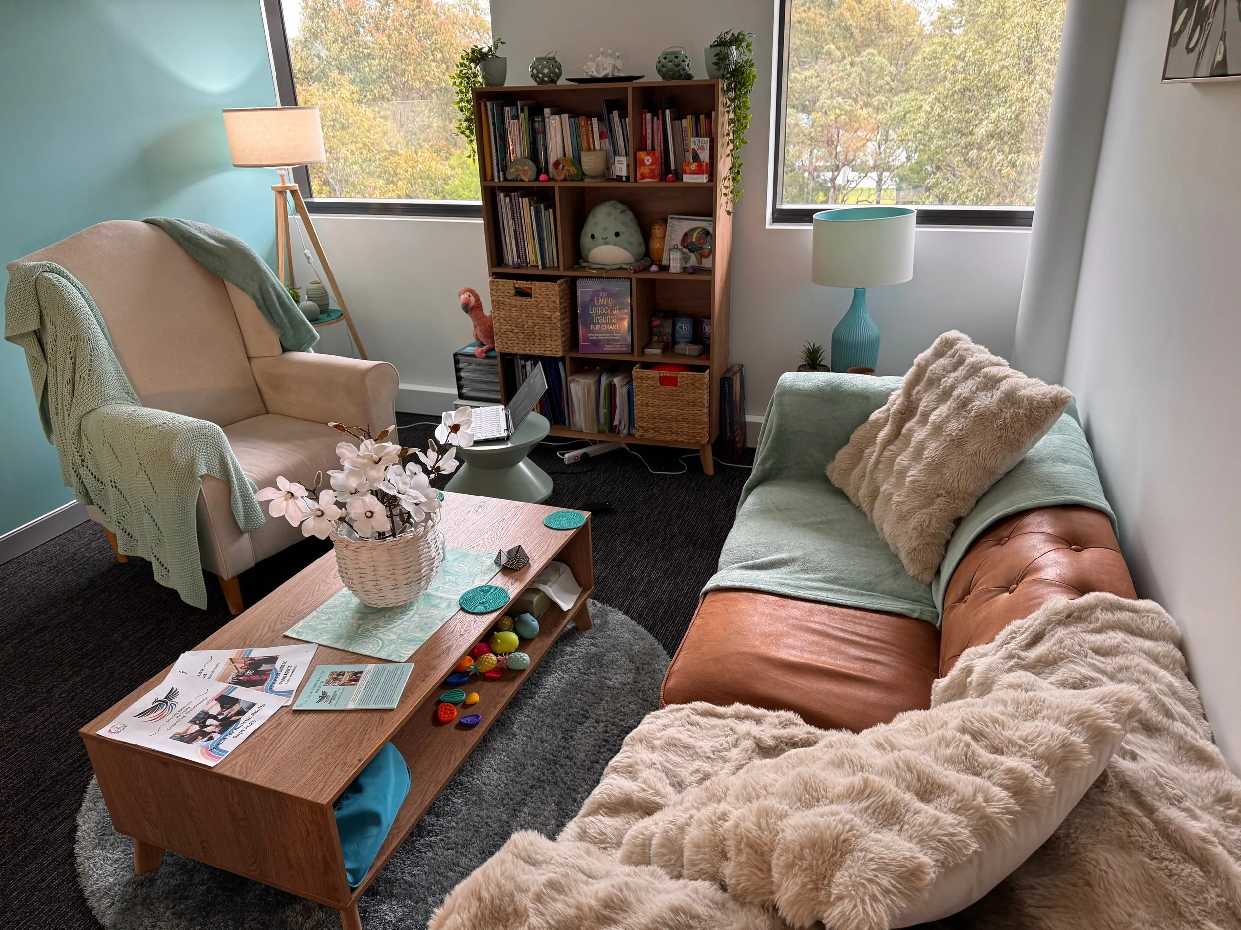 A cozy living room with large windows showing trees outside. Contains a beige armchair with a blanket, a green sofa with a fluffy blanket, a wooden coffee table with magazines and colorful toy pieces, a tall bookshelf with books and decorative items, and lamps providing warm lighting.