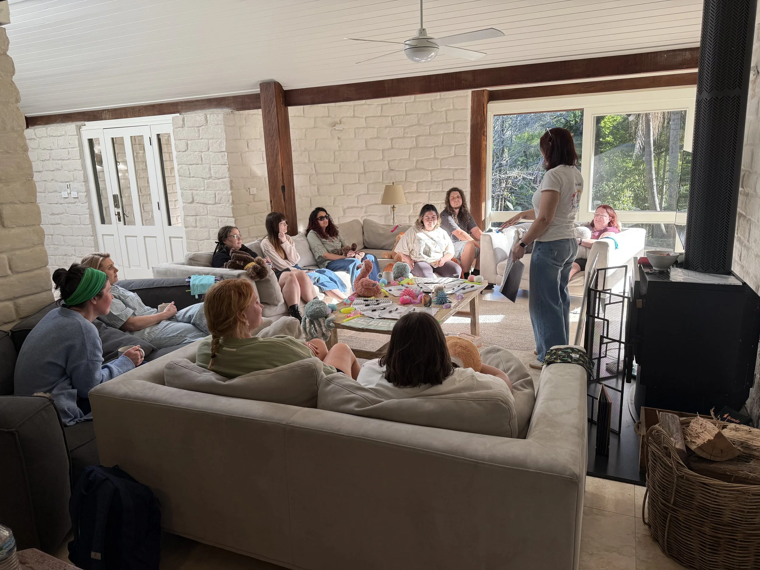 A group of women sitting on sofas in a living room, facing a woman standing and speaking. The room has large windows with trees outside, brick and wooden interior walls, and a ceiling fan.