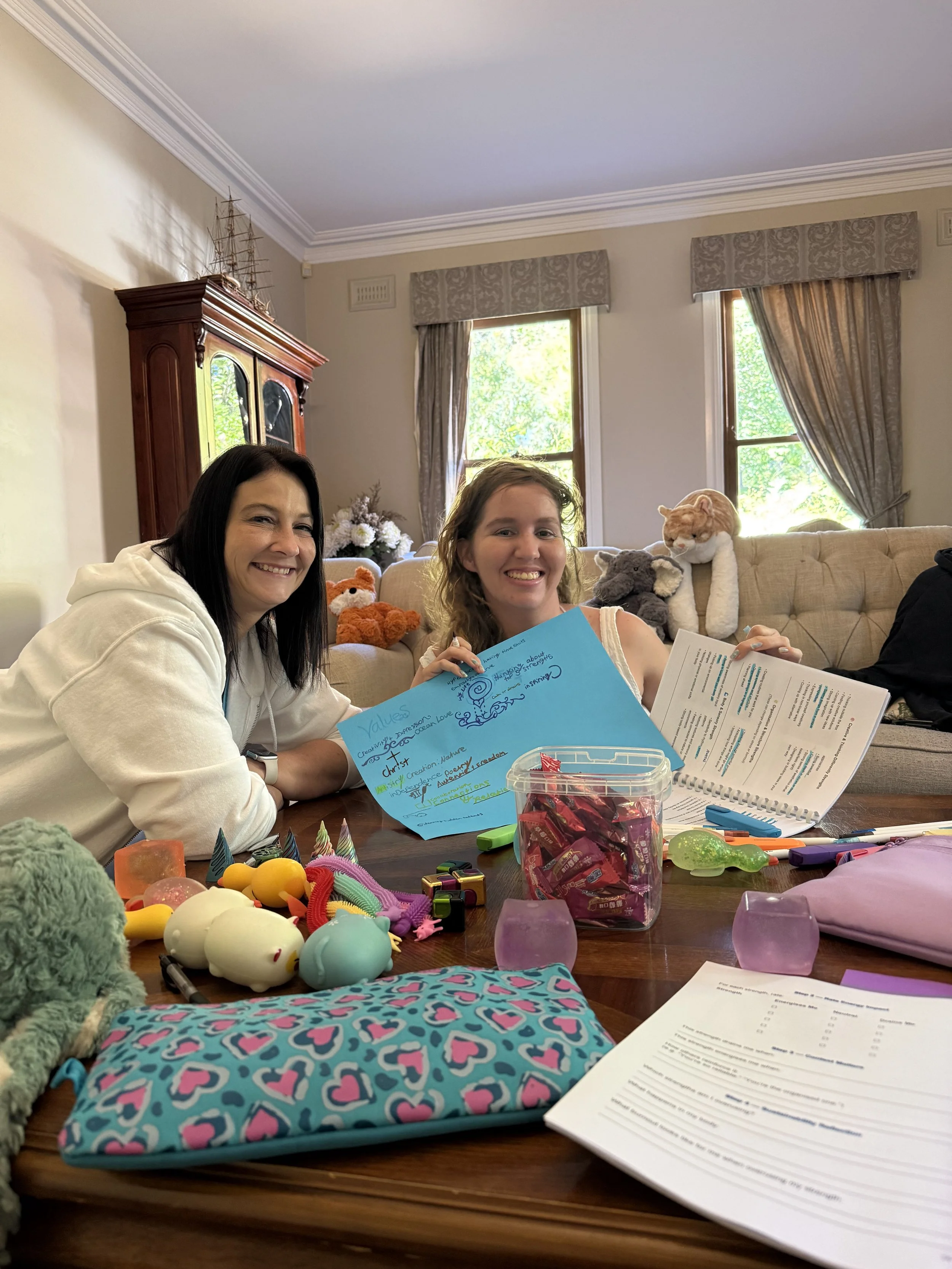 Two women sit at a table cluttered with toys, papers, and a blue paper with handwritten notes. Behind them, plush animals including a gray elephant, an orange cat, and a white bear are on a cream-colored sofa. The room has two windows with beige curtains and greenery outside. A wooden cabinet with model ships and a floral arrangement are visible in the background.