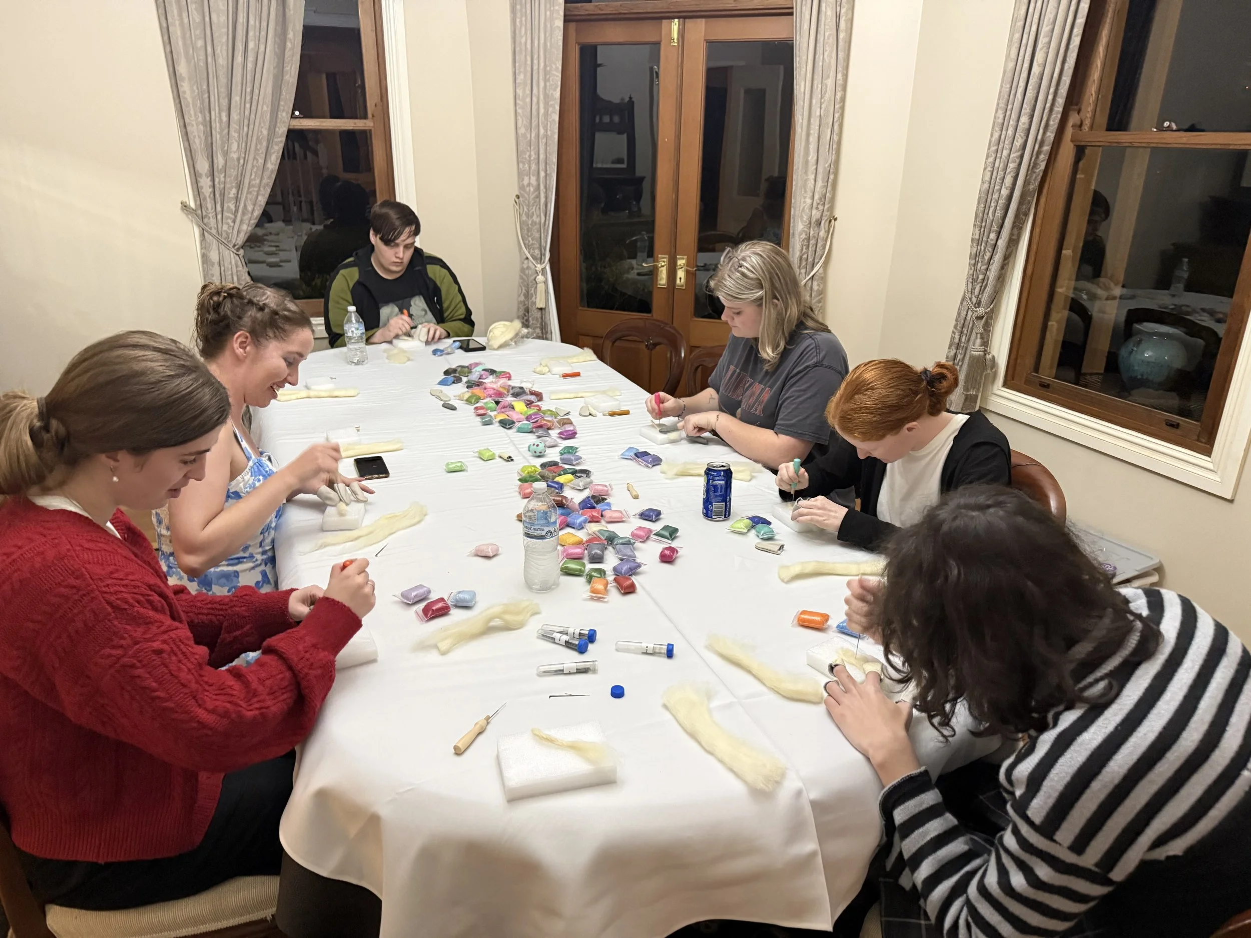 Group of people sitting around a table covered with various craft supplies, including fabric, scissors, and colorful decorations, engaged in a crafting activity in a cozy room with large windows and curtains.