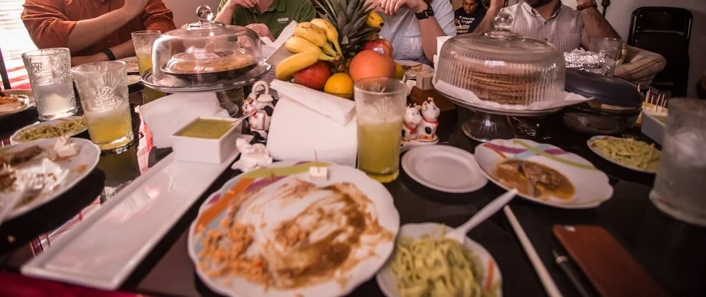 A party table with empty plates, partially eaten food, fruits, drinks, and cake, surrounded by people.