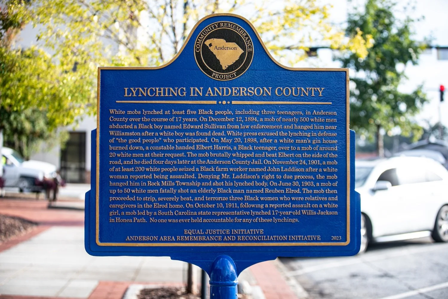 Blue historical marker plaque with gold border and black text, discussing lynching incidents in Anderson County, South Carolina, with a map icon at the top and trees and parked cars in the background.