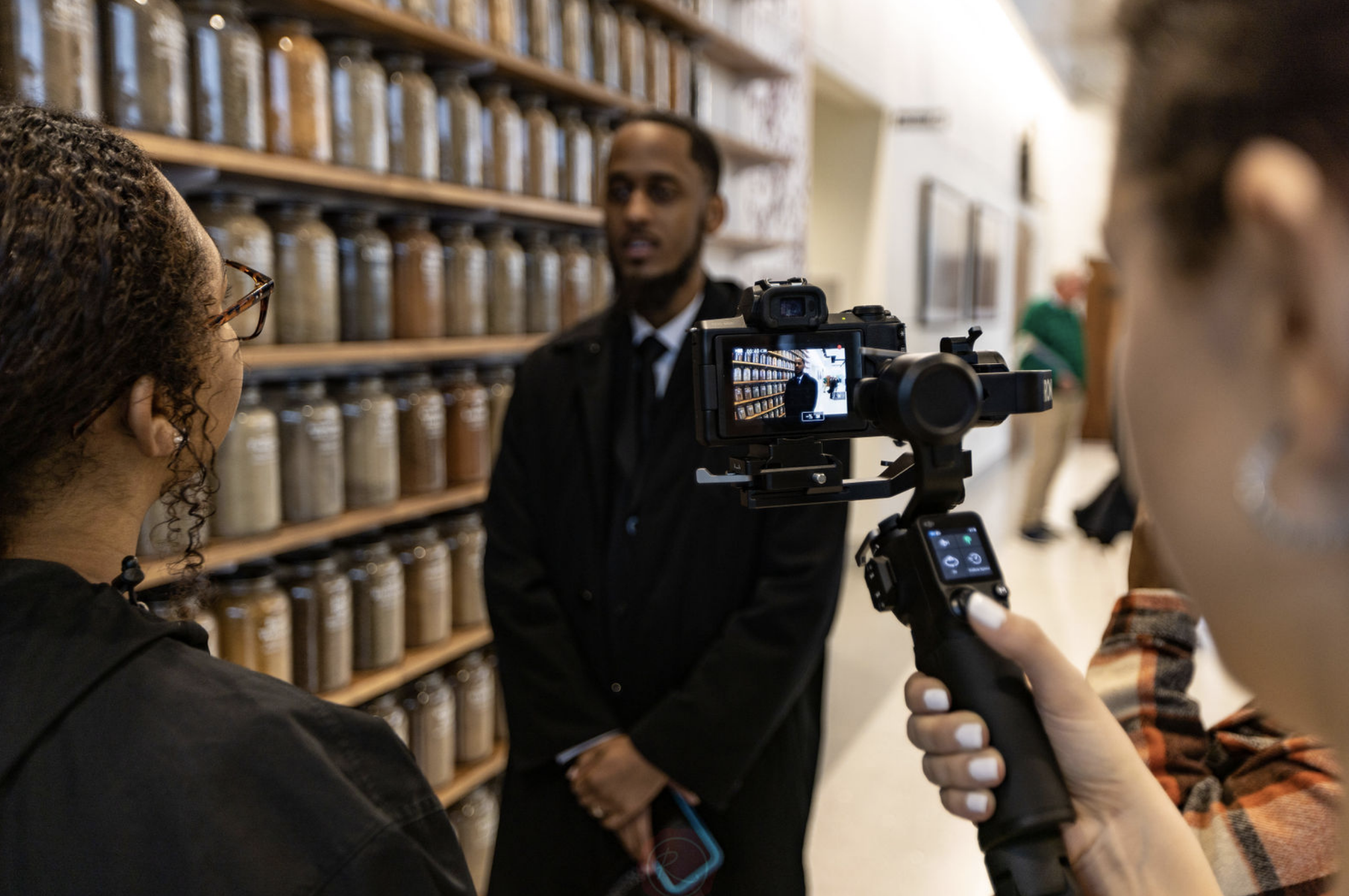 People filming an interview in a library or bookstore with shelves of jars or bottles in the background.