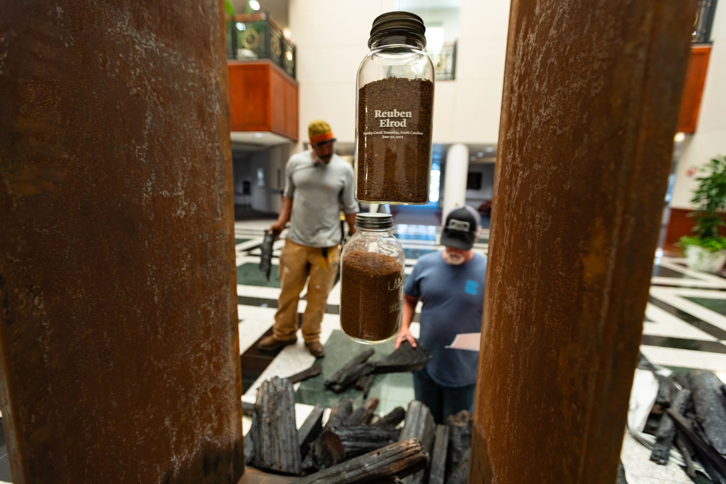 Close-up of a glass jar labeled 'Reuben Elrod' filled with soil, viewed from between two rusted metal beams, with two men in casual clothing and hats in the background at an indoor setting.