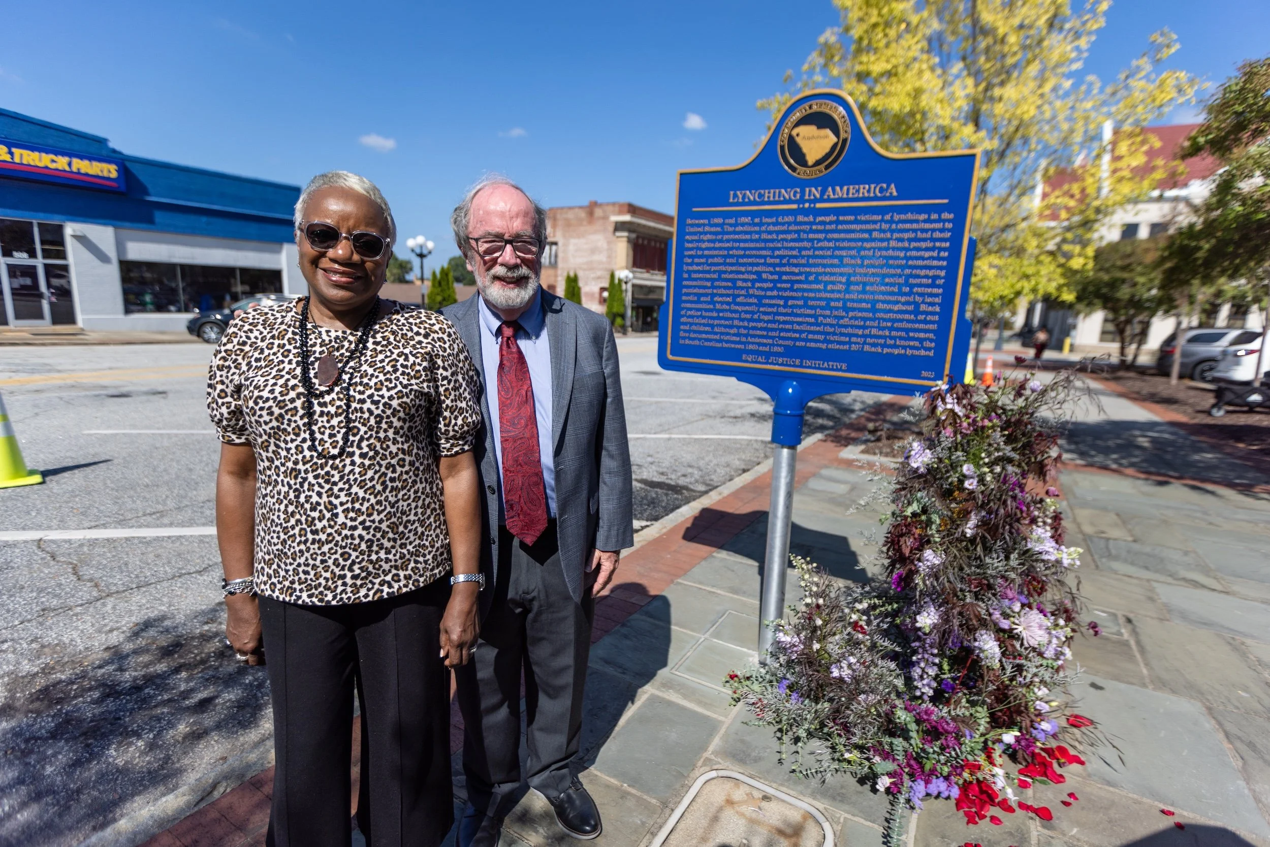 Two older adults, a woman with short gray hair wearing sunglasses and a leopard print top, and a man with glasses and a beard in a suit, stand together outside near a blue historical marker sign titled 'Lynching in America'. The sign provides information about racial violence against Black people in the U.S. The background includes trees, a building, parked cars, and a basket of flowers beside the sign.