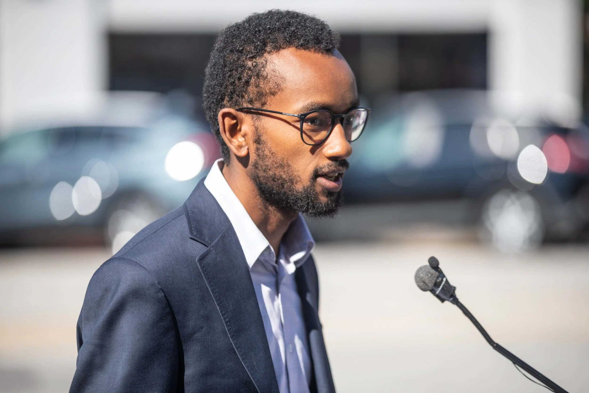 A man with a beard and glasses in a suit speaking at a microphone during a public event or press conference.