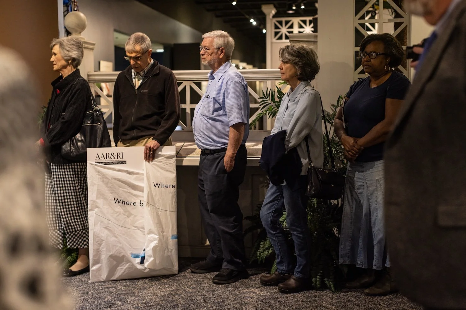 Six people standing in a row, some with their eyes closed or looking down, in a somber setting inside a building, participating in a memorial or remembrance event.