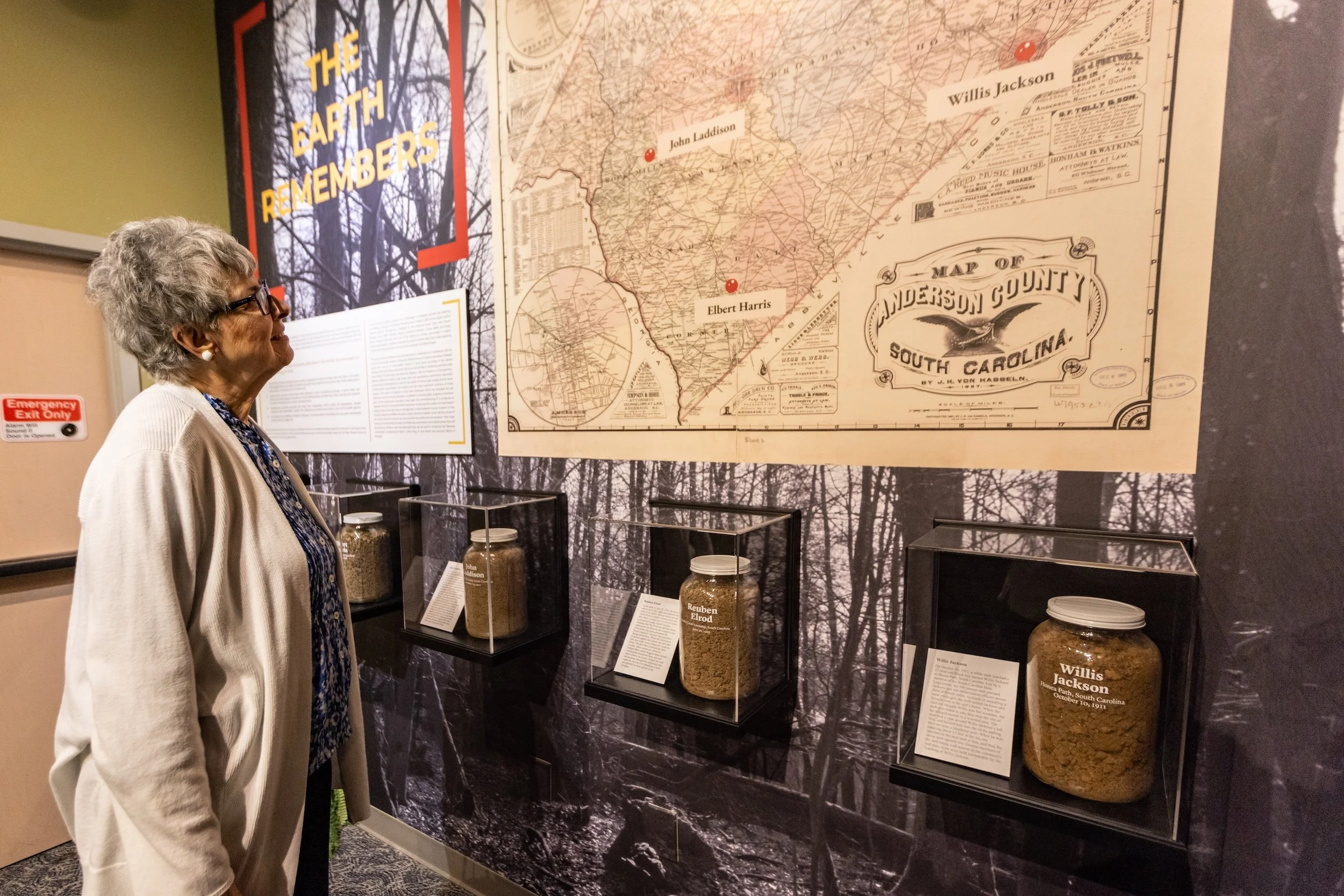 A woman with gray hair and glasses looking at historical display in a museum, featuring a large map of Anderson County, South Carolina, and display jars of grains with descriptive labels.