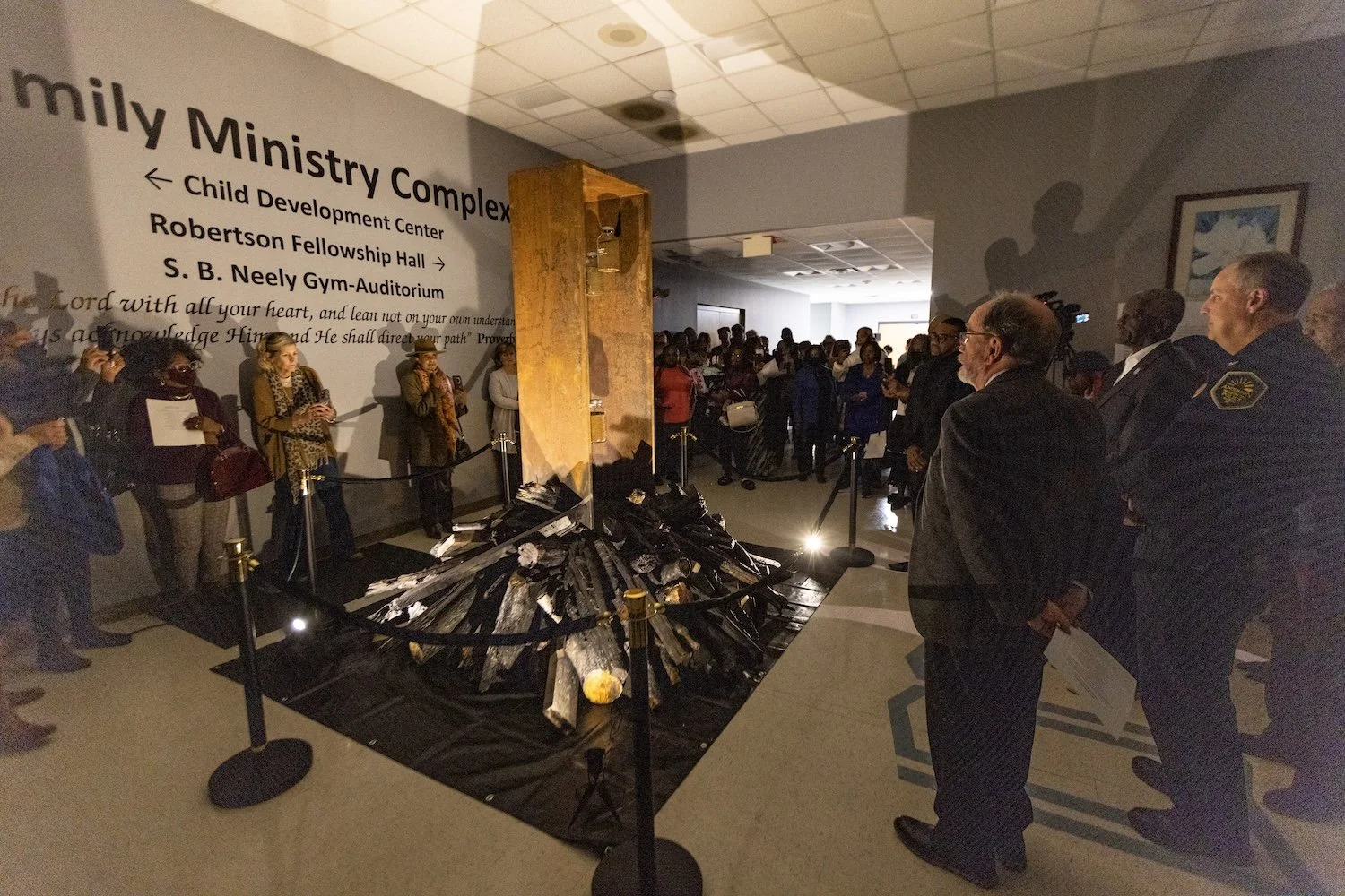A memorial display in a lobby with numerous people gathered around. The display features a pile of charred wood with a church pew behind it, roped off with black barriers. A large wall behind has text that reads 'Family Ministry Complex' and directions to various facilities, with a quote from the Bible.