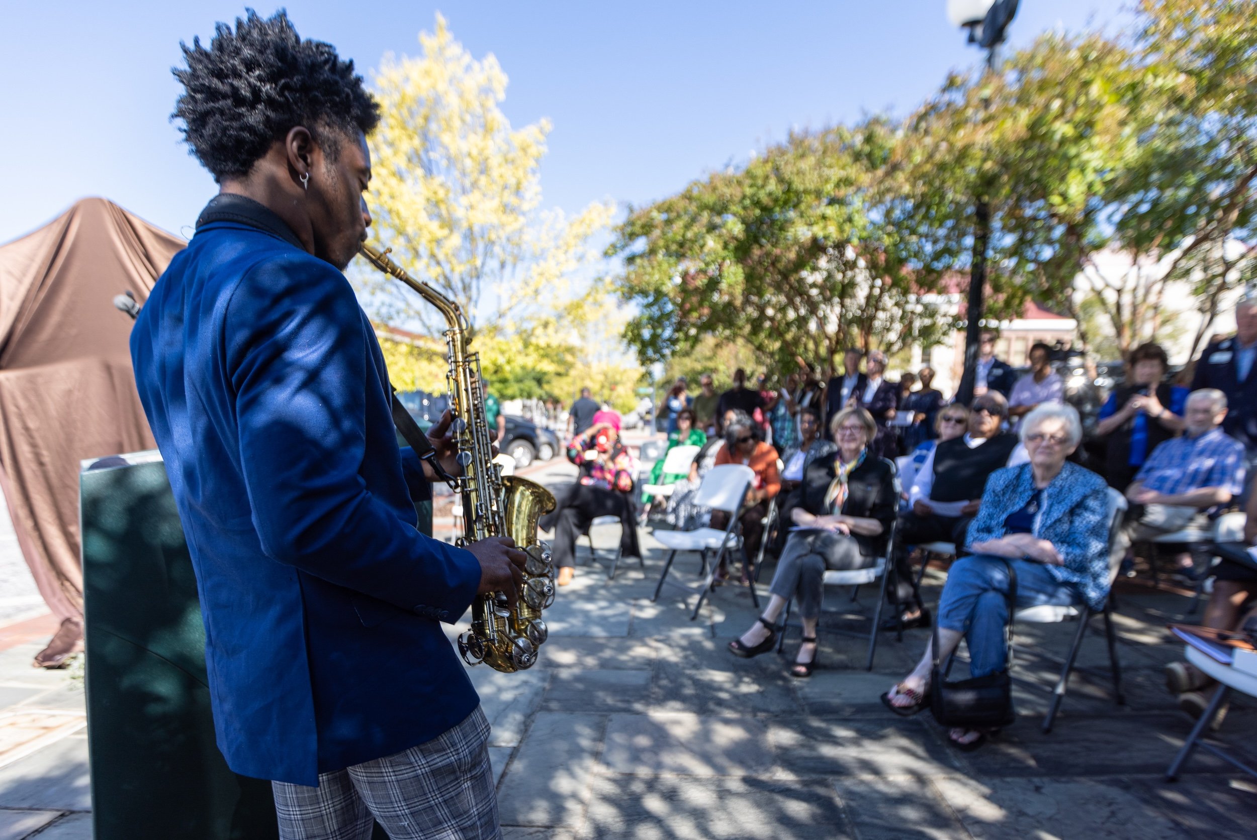 A man playing the saxophone outdoors during a public event or concert, with an audience seated on chairs in the background amid trees and blue sky.