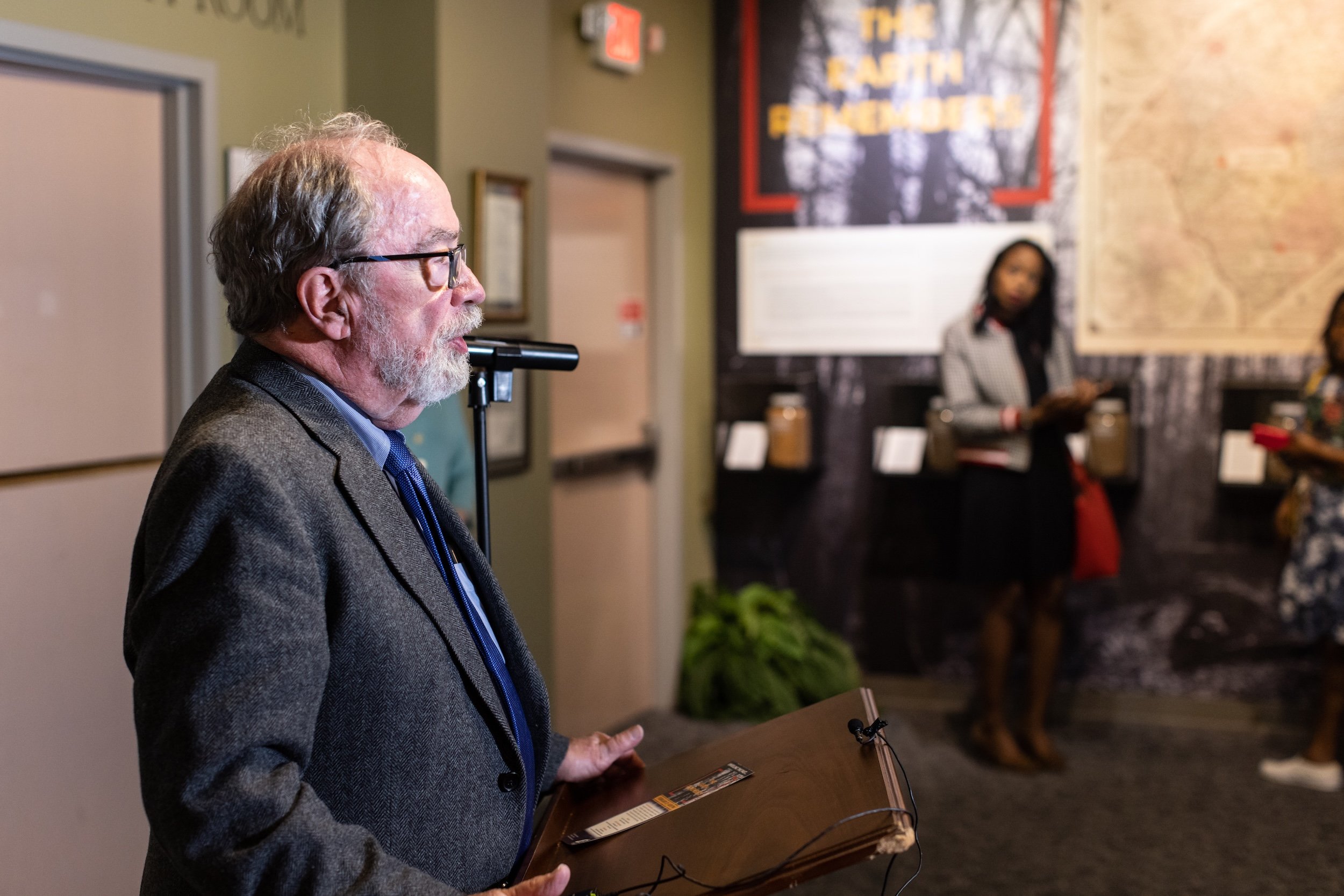 An older man with glasses and a beard wearing a gray jacket and blue shirt, standing inside a room, holding a book or brochure. In the background, a woman in a gray checkered blazer is standing near a wall with a map and decorative items.