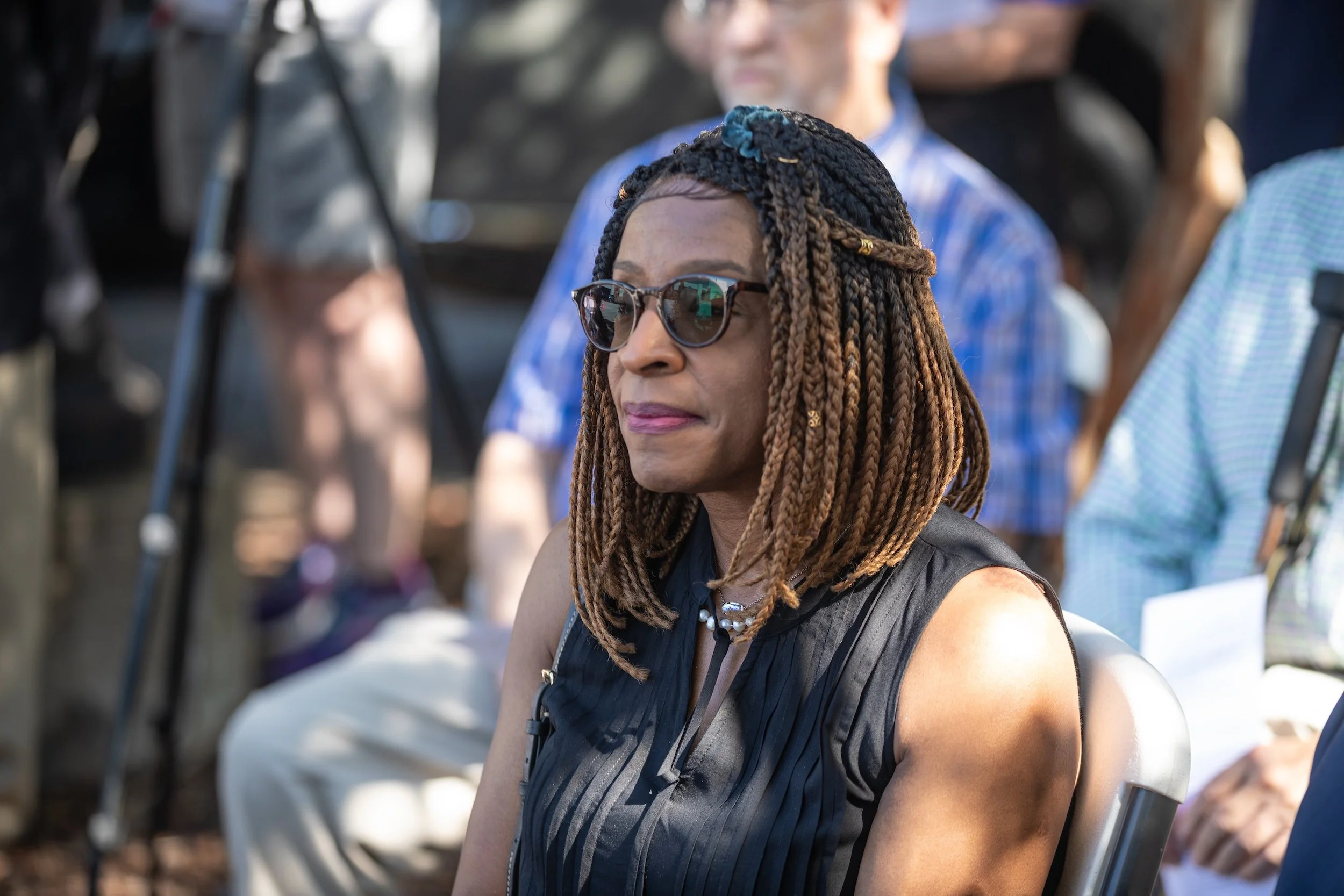 Woman with braided hair, sunglasses, and a black sleeveless top sitting outdoors, with another person visible in the background.