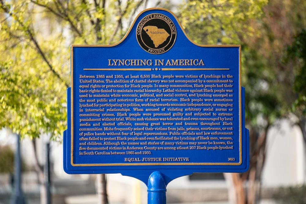 Blue informational sign about lynching in America in Anderson County, South Carolina, with a black and gold emblem at the top.